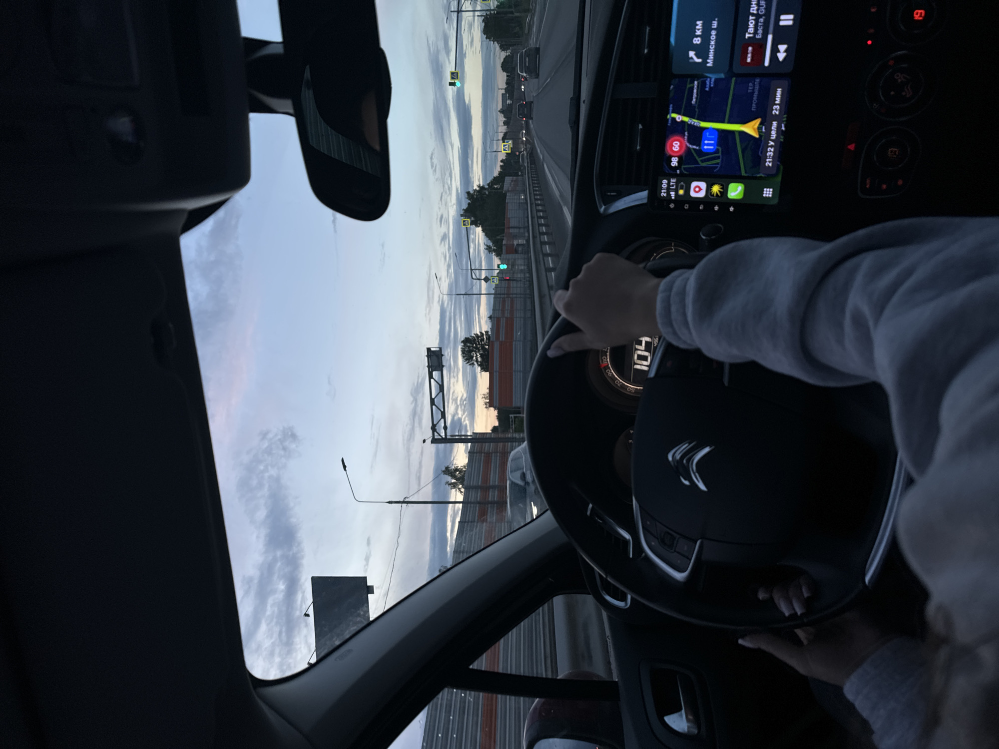 The interior of a moving Citroen vehicle is captured during twilight, heading toward Selyatino, Russia. The driver, wearing a light-grey long-sleeved shirt, has both hands on the steering wheel. The speedometer reads 104 km/h. The navigation system displays a route toward the destination. The estimated time of arrival is 21:32, and the remaining travel time is 23 minutes. The route is indicated on the map, and the current location is 8 kilometers from the destination, which is along the Minskoye Highway. Exterior elements visible through the windshield include road signage, street lights, and the sky.