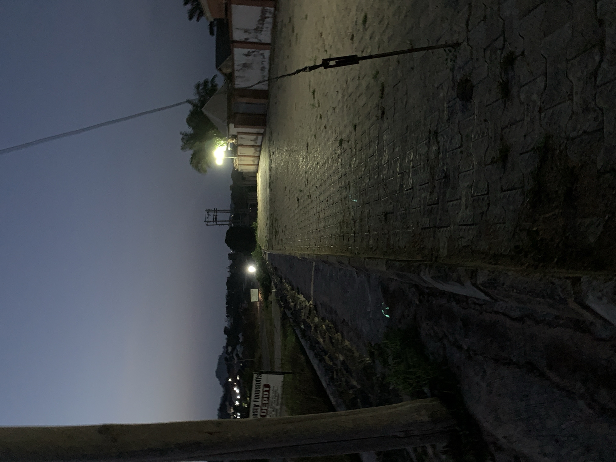 The image depicts a twilight scene on a paved walkway in Gbeleko, Nigeria. A wooden pole stands to the left of the frame, with a sign reading "nasty Foodstuffs DEPOT" visible behind it. The walkway is illuminated by streetlights, casting long shadows and highlighting the texture of the paving stones. A low stone wall runs along the left side of the walkway. In the background, buildings and a distant hill are visible, with scattered lights suggesting a developing urban area. A thin wire stretches diagonally across the upper right portion of the image. The overall impression is one of a quiet evening in a developing neighborhood.