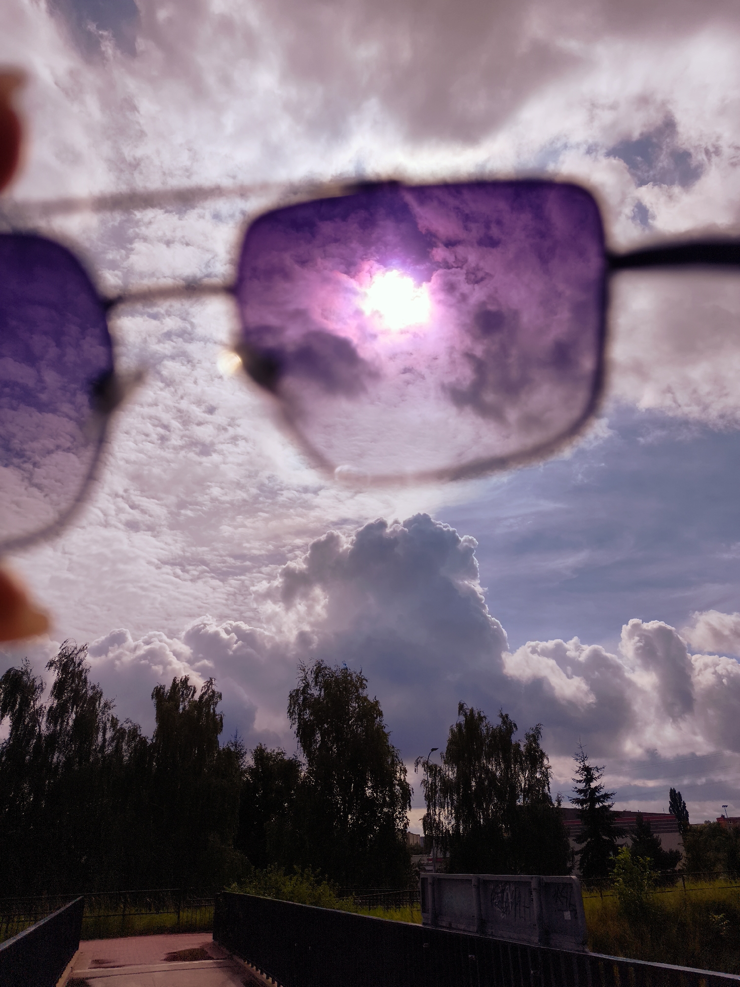 The image shows a close-up of a pair of sunglasses reflecting a cloudy sky with the sun shining brightly. The sunglasses lenses are tinted purple. In the background, out of focus, is a line of trees and a glimpse of buildings. The lower part of the image shows a bridge or walkway with railings. The scene suggests an outdoor environment, possibly a park or a street with vegetation and a bridge. The presence of buildings in the distance indicates an urban or suburban setting. The overall lighting suggests daytime, with the sun being a prominent element in the reflection.