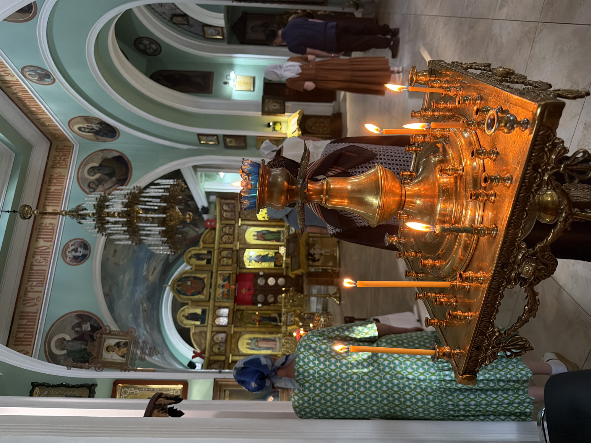 The image depicts the interior of a church, likely an Orthodox Christian church, given the iconography and altar screen visible in the background. In the foreground, a large, ornate golden candelabrum holds several lit candles, casting a warm glow. A woman in a green and white patterned dress and a blue headscarf stands with her back to the camera, facing the altar. Other individuals, including a woman in a brown dress and headscarf and a man in a dark blue shirt and pants, are also present, facing the altar area. The church features high ceilings, arched doorways, and numerous religious icons painted on the walls and ceiling. The altar area is elaborate, with a large iconostasis adorned with multiple icons and decorative elements. The overall atmosphere appears to be one of quiet reverence. Based on visual cues, the style of the church is consistent with Orthodox churches found in Russia, and the presence of iconography and liturgical items further supports this. Without specific external landmarks or signage within the image, pinpointing the exact church in Sochi, Russia, is not possible from the visual information provided.
