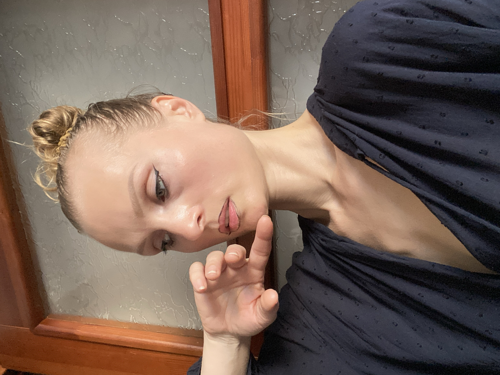 A close-up, chest-up studio portrait of a young woman with fair skin and blonde hair pulled into a bun is shown. She is wearing a dark blue, textured blouse. Her left hand is raised to her chin, with fingers bent and touching her lips. Her head is tilted to her left, and her gaze is directed downward. The background consists of a wooden frame and a textured wall with reflective elements. There is no indication of the city of Lida, Belarus.