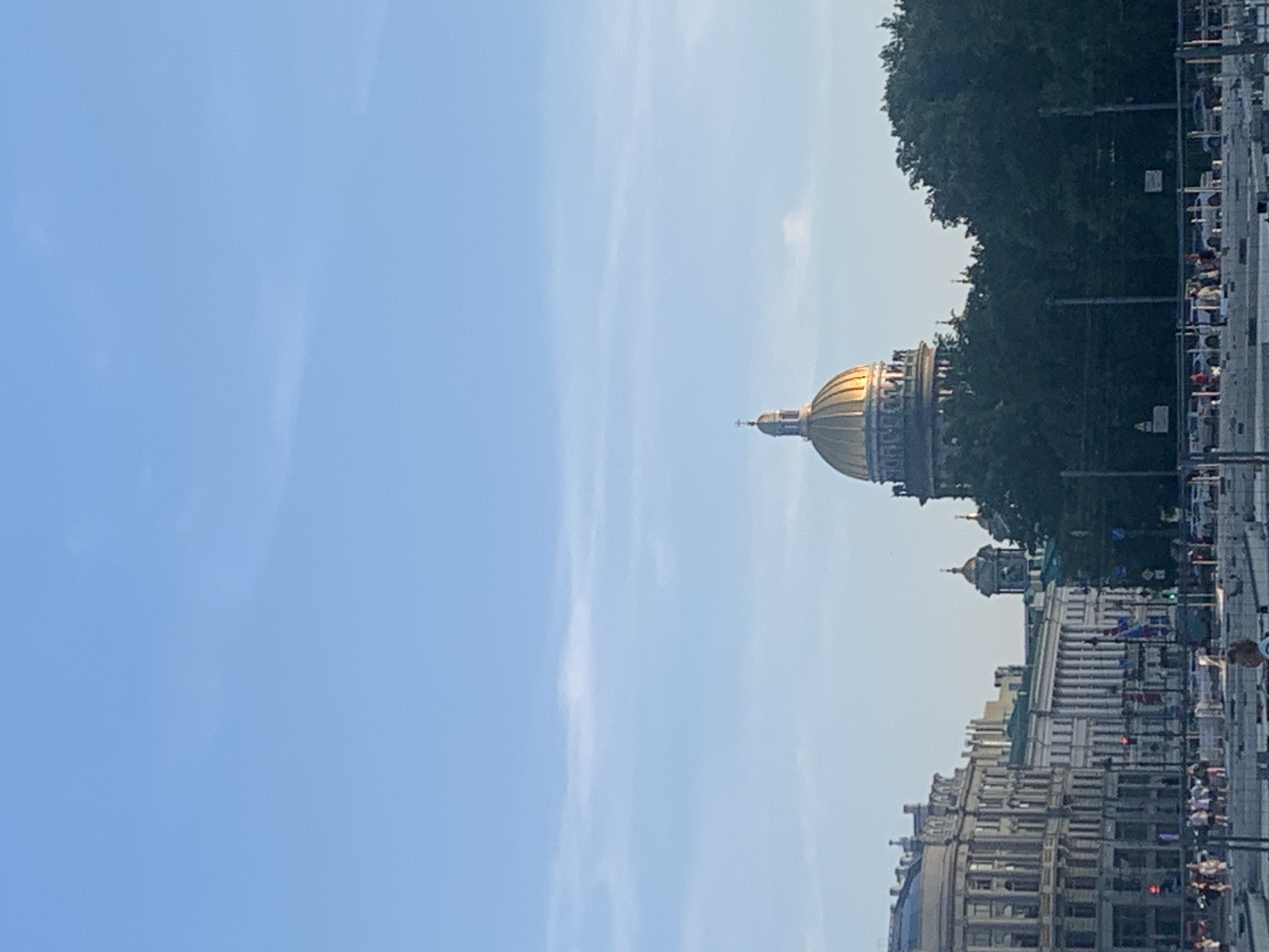 The image captures a view of Saint Isaac's Cathedral in Saint Petersburg, Russia, with its iconic golden dome prominently featured against a clear blue sky with sparse clouds. In the foreground, a row of neoclassical buildings lines a street. Trees obscure the lower portion of the cathedral's dome. The scene appears to be set in a public area, possibly a square or embankment, with several pedestrians visible in the mid-ground. There are also vehicles present on the street. The overall impression is of a sunny day in an urban environment.