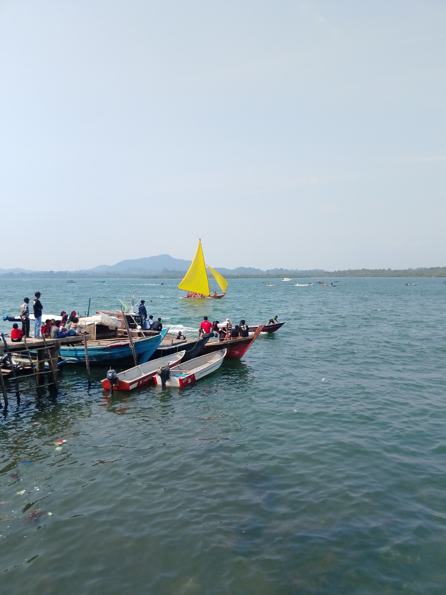 The image depicts a waterfront scene in Batam, Indonesia, likely at a jetty or harbor. Multiple boats are visible, ranging from small motorboats to larger fishing vessels. A distinct yellow sailboat with a single sail is prominent in the mid-ground, heading towards the right of the frame, carrying several passengers. On the left, a wooden jetty is occupied by a group of people, some standing and observing the water, while others are seated. Several other boats are moored alongside the jetty, with people on board or disembarking. The water is choppy, indicating a moderate breeze. In the background, a lush green coastline with rolling hills forms the horizon under a pale sky. The presence of debris in the water near the jetty suggests potential pollution.