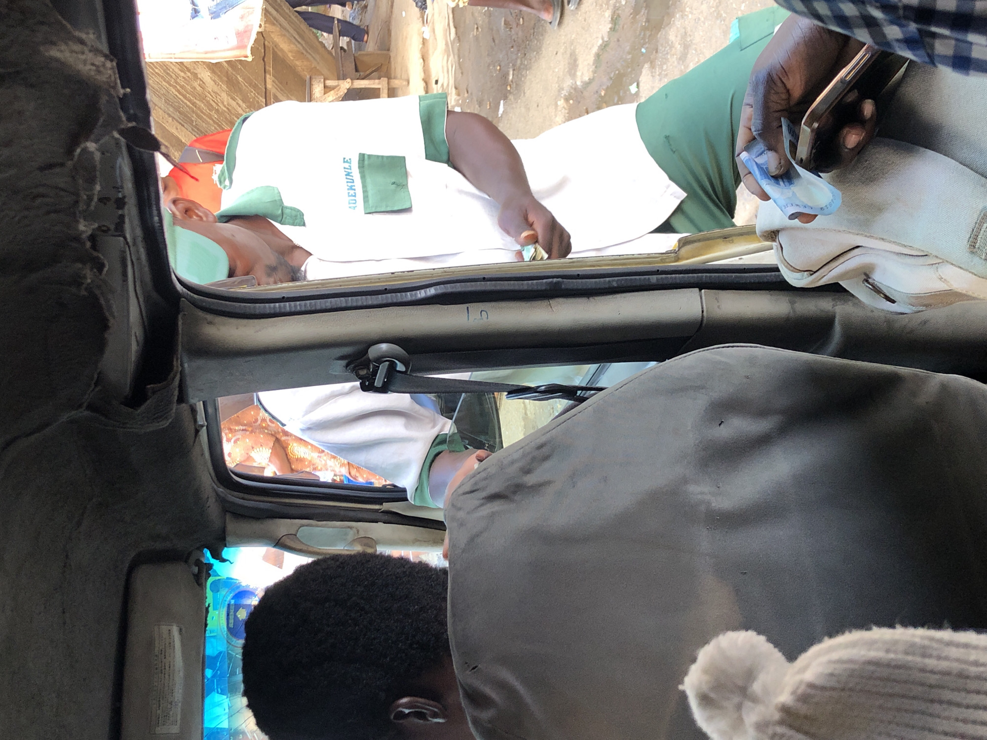 The image captures a scene inside a vehicle, likely a bus or taxi, in Ikeja, Nigeria. The foreground shows the back of a person with dark, curly hair and the shoulder of a person wearing a dark grey garment. A light-colored knitted hat with a pom-pom is visible in the lower left corner.
Through the open doorway, a person wearing a white shirt with green trim and a green cap is seen extending their hand, holding what appears to be cash. The shirt has the embroidered name "ADEKUNLE" on the chest. Their body is angled towards the inside of the vehicle. In the background, other individuals and what seems to be a market or roadside environment are visible, with some stalls and a wooden structure. The ground is dusty and appears somewhat wet in places. The overall impression is of a busy, urban, and possibly commercial setting within Ikeja.