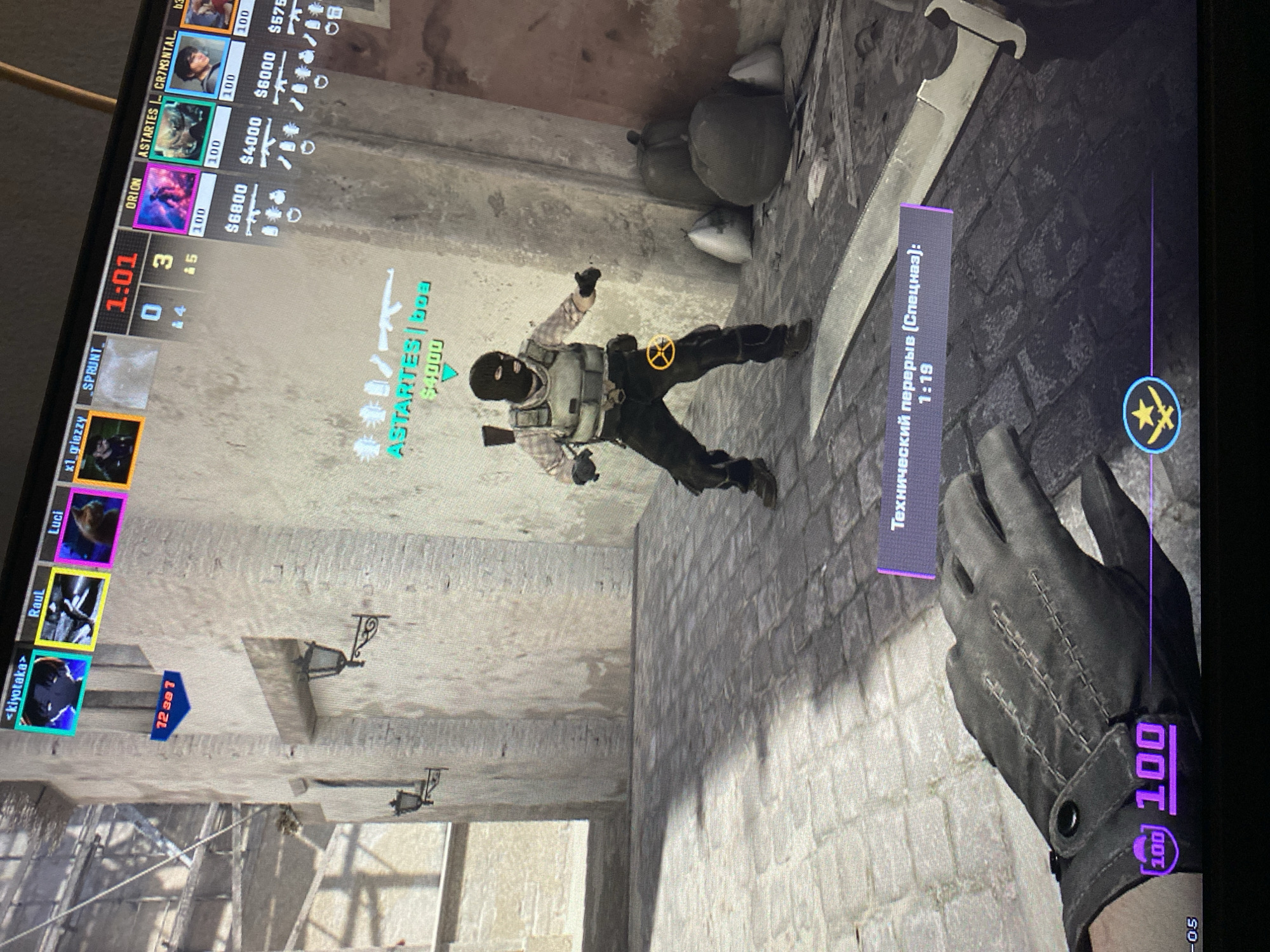 The image displays a screenshot from a first-person shooter video game, likely Counter-Strike, seen from the player's perspective. The scene depicts an in-game character, resembling a terrorist or insurgent with a balaclava, tactical vest, and a rifle slung on their back, standing against a concrete wall. Behind the character are several sandbags or sacks.
In the foreground, the player's gloved hand and wrist are visible, along with a large combat knife. The game's user interface is prominent, featuring a scoreboard on the left with player names such as "x1_griezzy," "kiyotaka," "Raul," "Luci," "ORION," "ASTARTES," and "CR7M3NTAL," along with their respective in-game money and weapon icons. A timer at the top-left shows "1:01," and scores "0" to "3" are displayed. The character in the center also has an "ASTARTES" tag above their head with "$4000."
A significant detail is a purple text box on the right that reads "Технический перерыв (Спецназ): 1:19," translating to "Technical pause (Special Forces): 1:19," indicating the game is currently paused. The player's health or armor is displayed as "100" in the bottom right corner. The game environment appears to be an urban or industrial setting, characterized by concrete and brick walls, with some structural elements and wall-mounted lights in the background. While the context of Yerevan, Armenia, is provided, the image itself shows a virtual game world and offers no specific real-world geographical cues.