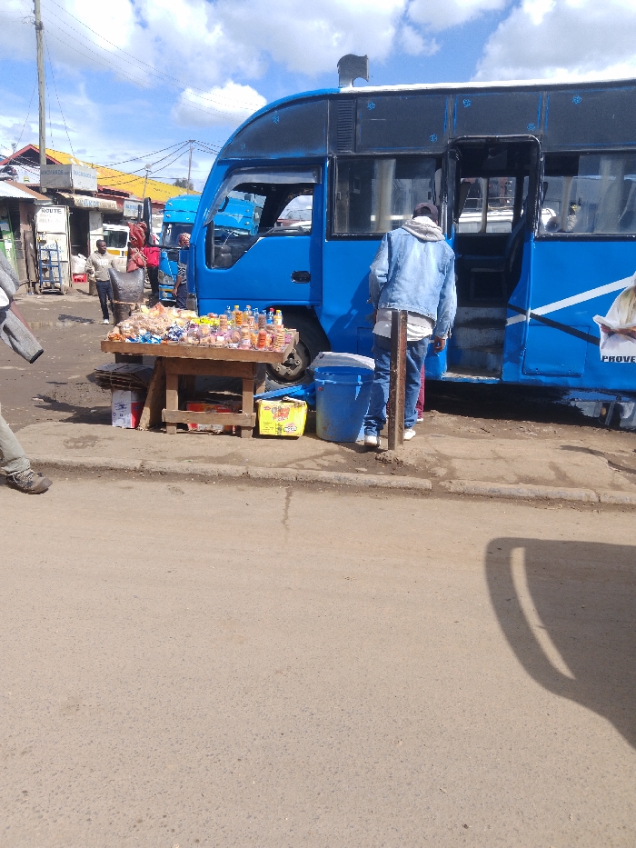 The scene is a street corner in Athi River, Kenya, near the coordinates -1.453 latitude and 37.007 longitude. A blue bus is parked along the curb with its doors open. A man in a blue denim jacket is standing near the bus with his back to the camera, looking into the bus. A wooden table is in front of the bus. The table is covered with various items including what appear to be snacks and small bottles of liquid. Another man is standing to the left of the table, looking towards the street. The street is dirt and is lined with a curb. The bus is brightly painted in a light blue color. The sky is partly cloudy.