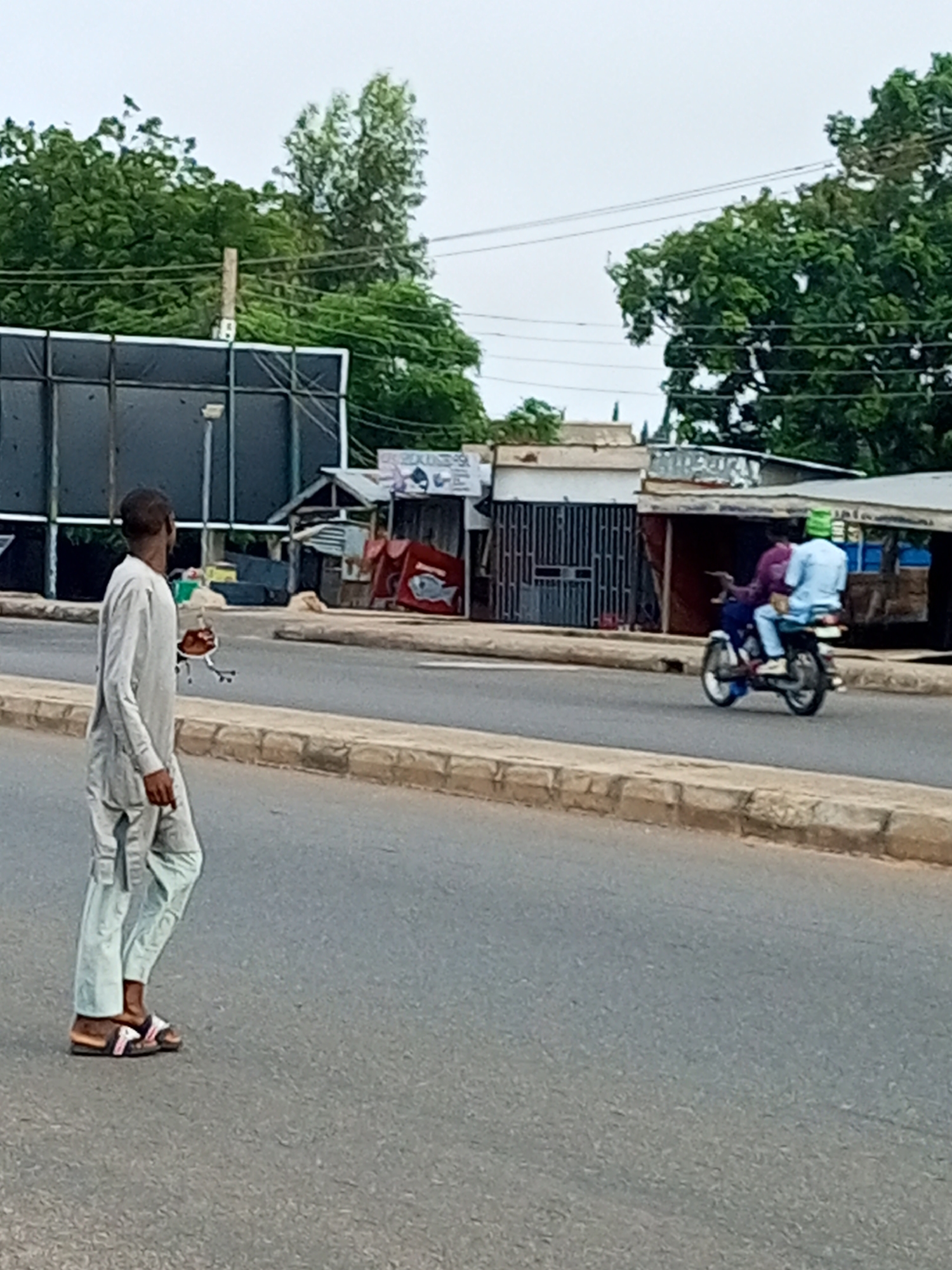 In Garko, Nigeria, a man in a light-colored traditional outfit walks on a paved road next to a curb. He is looking towards the left. In the background, two people are riding on a motorcycle. The person in front is wearing a purple shirt and the person behind is wearing a light blue shirt and a green cap. They are traveling on the same road as the pedestrian. Behind the road, there are several small buildings with corrugated metal roofs and some foliage. A large dark billboard is visible on the left side of the image. The sky is overcast.