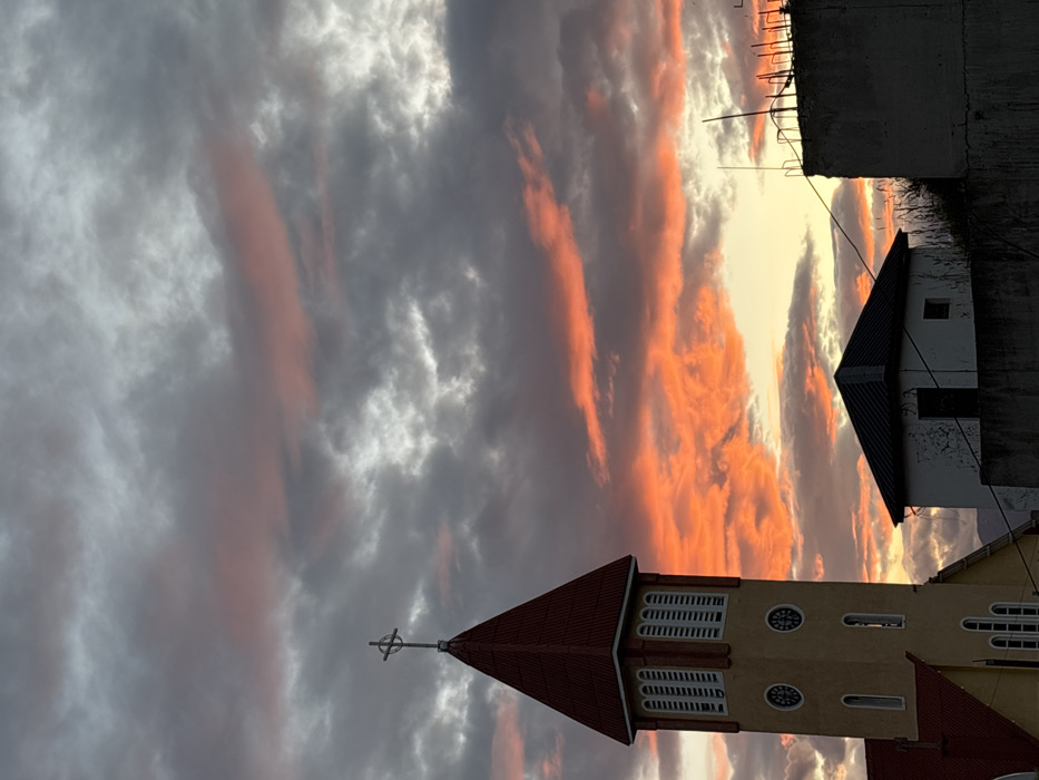 A church steeple with a cross is silhouetted against a dramatic sky filled with gray and orange clouds. Adjacent to the church is a building with a black roof, and a partial view of a concrete structure sits above it. The sunset creates a striking contrast between the dark buildings and the vibrant sky. The image is rotated.