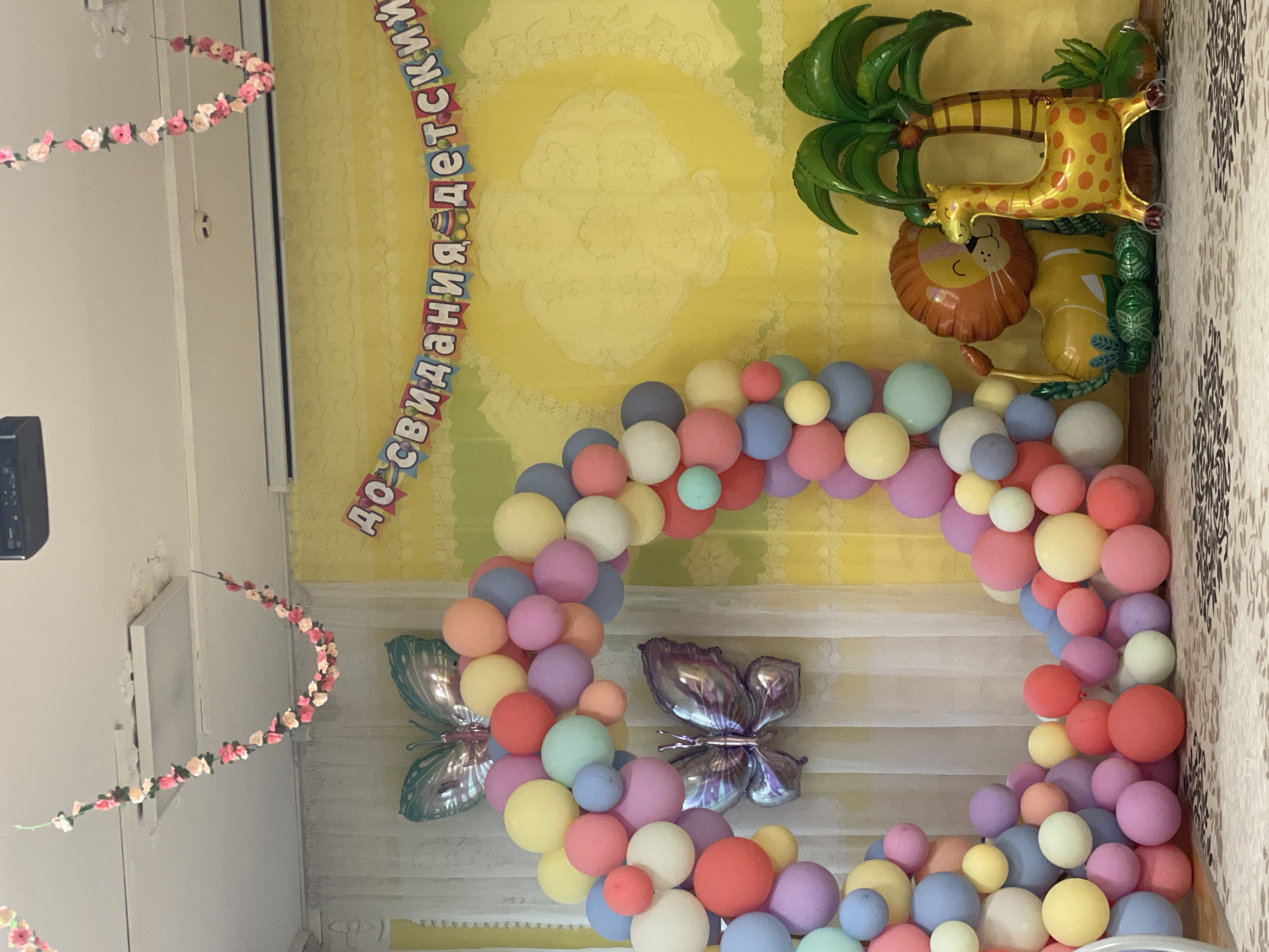 The image shows a room decorated for a celebration, likely a child's party, with a balloon arch in pastel colors, butterfly-shaped foil balloons, and an inflatable jungle scene with a lion, giraffe, and palm tree. There is a banner with the text "до свидания детский сад", which translates to "goodbye kindergarten" in Russian. The room appears to be in Izhevsk, Russia, given the Russian text. Additionally, flower garland is present on the ceiling.