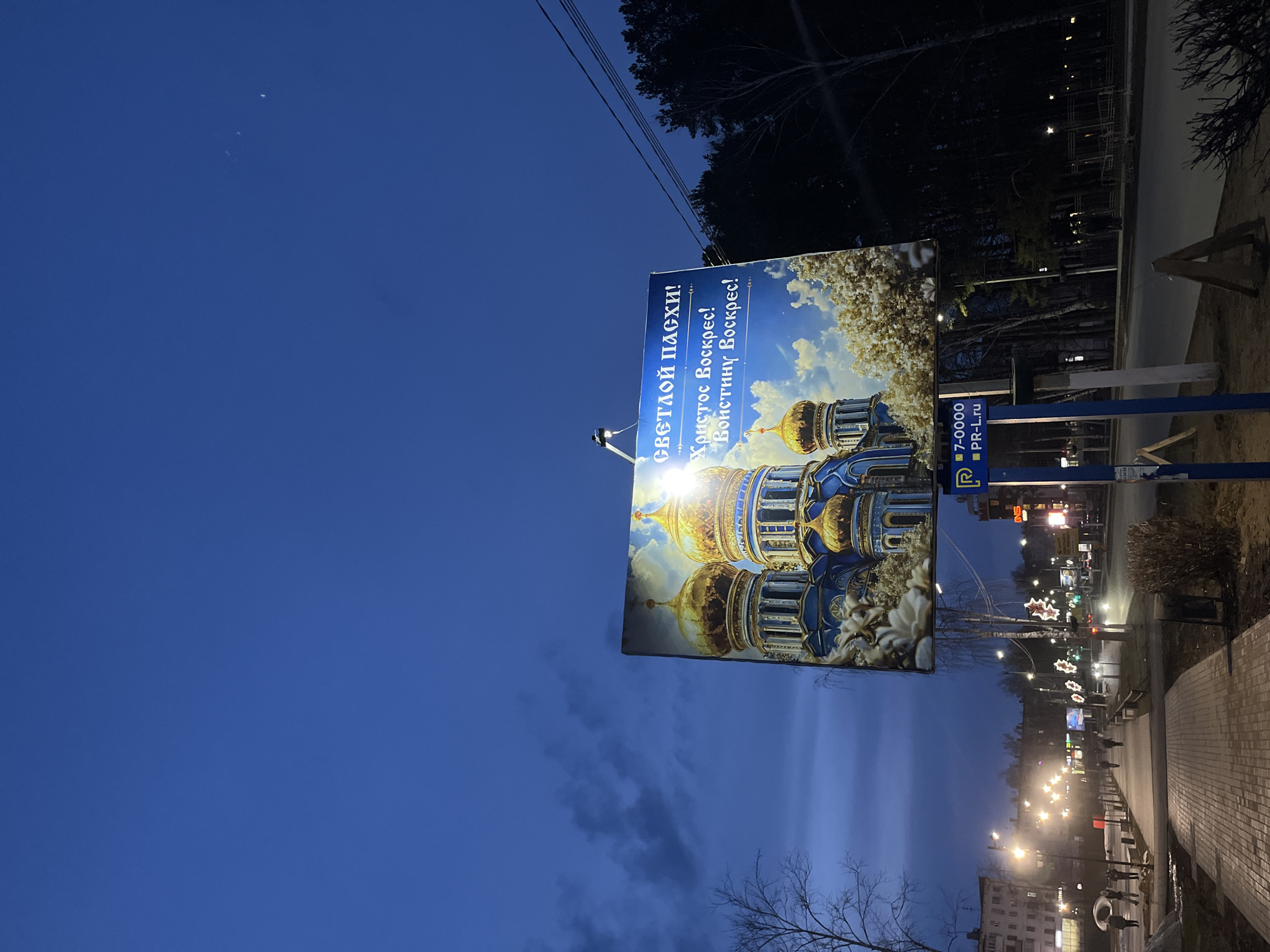 The image shows a billboard in the city of Chaikovsky, Russia. The billboard has the text “Светлой Пасхи! Христос Воскрес! Воистину Воскрес!”, meaning “Happy Easter! Christ is Risen! Indeed He Is Risen!”. The billboard also features an image of a Russian Orthodox church. Below the billboard, there is a sign with the phone number 7-0000 and the website pr-l.ru. In the background, there is a street scene with streetlights and buildings. The sky is blue with some clouds. There is a sidewalk in the foreground. The lighting suggests it is evening or early morning.