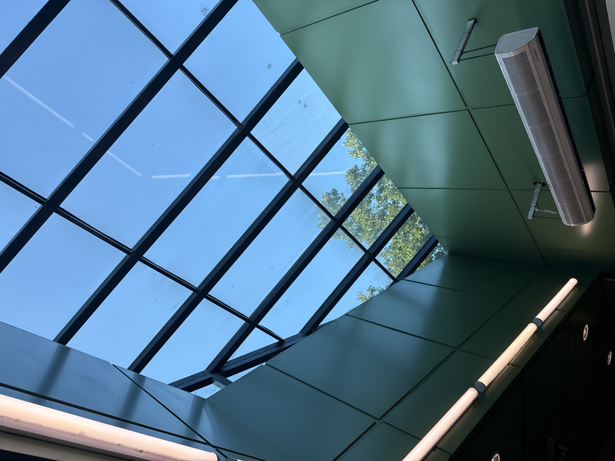 The image shows an interior view looking up towards a modern skylight. The skylight is composed of glass panes framed by dark metal beams, creating a grid pattern. Through the glass, a clear blue sky and the green foliage of trees are visible. The interior walls are clad in large, green panels, and the ceiling features fluorescent lighting and what appears to be an air curtain unit mounted on the wall. The architectural style suggests a public space such as a shopping mall, transit station, or modern office building. The specific location in Sofia, Bulgaria cannot be determined from the visual information alone.