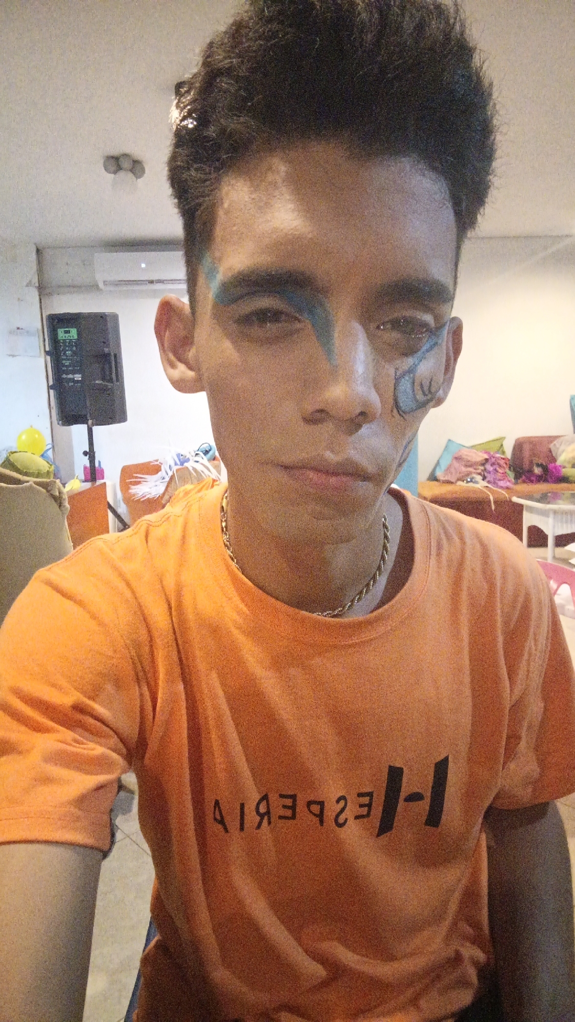 A young man poses for a selfie in an indoor setting, likely a residential area. He has dark hair, tan skin, and is wearing an orange T-shirt with a reversed or mirrored inscription in black. He has blue face paint extending from his left eyebrow and another design near his right eye. He is wearing a gold chain. The background includes a black speaker on a stand, a white air conditioning unit mounted on the wall, and furniture covered in blankets and pillows. A glimpse of a yellow balloon and other items suggests a casual, possibly festive environment. The location is identified as Playa El Agua, Venezuela, but specific landmarks are not visible.