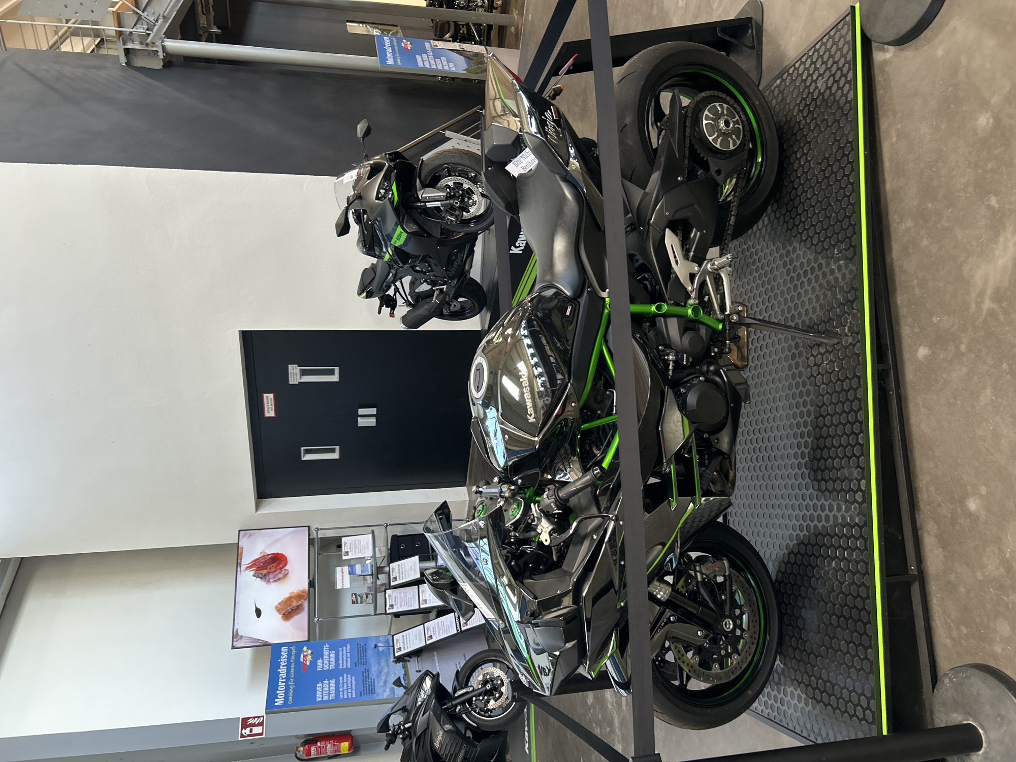 Here is a description of the image.
The image depicts a display of Kawasaki motorcycles, likely in a dealership or showroom. The foreground features a Kawasaki Ninja H2 with a black and green color scheme. It is displayed on a black platform with a neon green border. Behind it, another motorcycle, a Ninja, is visible. In the background, there are informational displays and signs. The image is taken indoors, possibly in Bernhausen, Germany, based on the writing on the signs that include the name Motorradreisen, and the German texts. The location appears to be well-lit, with a clean and organized environment.