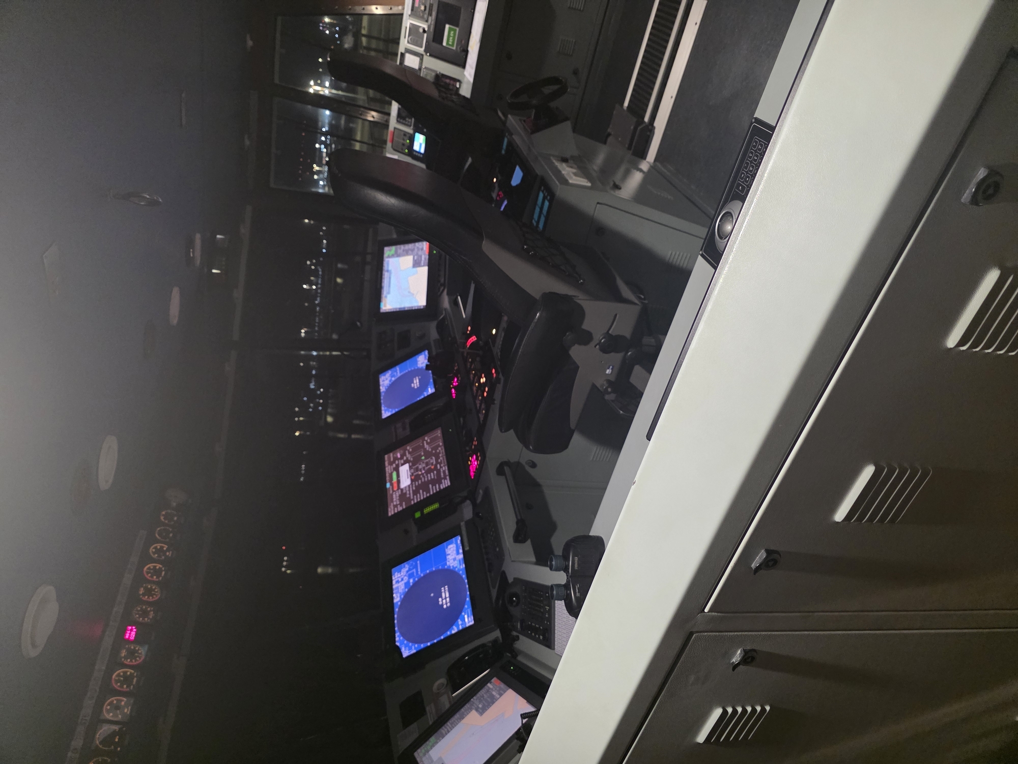 The image depicts the bridge of a ship at night, likely docked in the city of Brindisi, Italy. The dimly lit interior features a complex control console with multiple screens displaying navigational information and radar data. Two captain's chairs are positioned at the console, one of which is occupied by a person whose back is to the camera. Various buttons, switches, and a steering wheel are visible on the console. Outside the windows, the lights of a city and port are faintly visible against the dark night sky. The overall scene suggests a functional and operational vessel's command center.