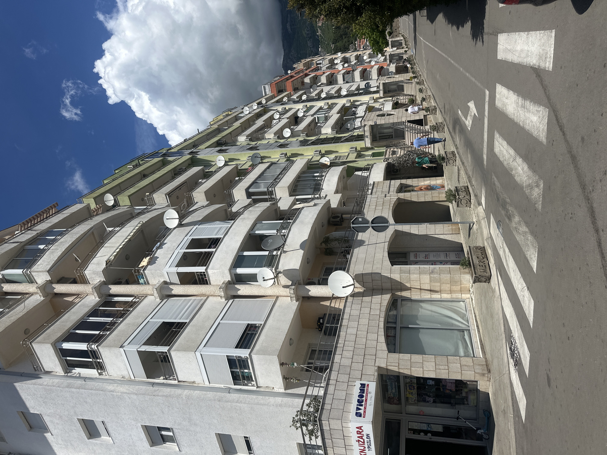 The image depicts a multi-story apartment building in Budva, Montenegro. The building has multiple balconies, most of which are equipped with satellite dishes. A street runs alongside the building, featuring a pedestrian crosswalk. A sign for "KNJIZARA YPSILON" is visible on the ground floor. A large cloud formation is present in the sky above and behind the building. Two individuals can be seen standing near the entrance to the building.