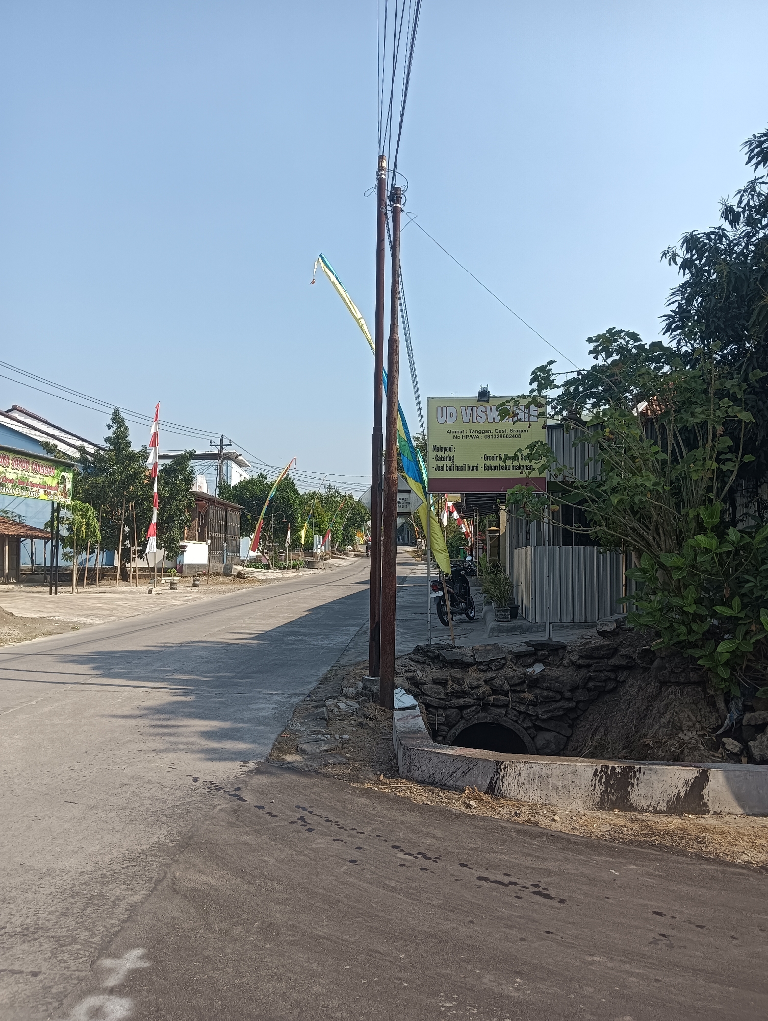 The image shows a street scene in the city of Tanggan, Indonesia. The road is paved and appears to be a two-lane street. On the left side of the road, there are several buildings, including what appears to be a store with a sign that reads "UD VISVA." There are also trees and red and white flags lining the street. On the right side of the road, there are more trees and some fencing. A motorcycle is parked on the right side of the road. The sky is clear and blue. The overall impression is of a quiet, rural or suburban street.