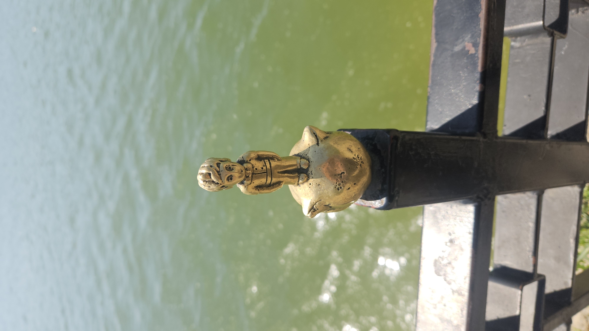 A golden statue atop a black iron railing is positioned with a body of water in the background. The statue appears to be a human figure standing on top of a sphere with animal features. The railing is part of a larger structure with multiple horizontal and vertical bars. The water in the background has a green hue. The location is likely Chișinău, Moldova, based on the statue's style.