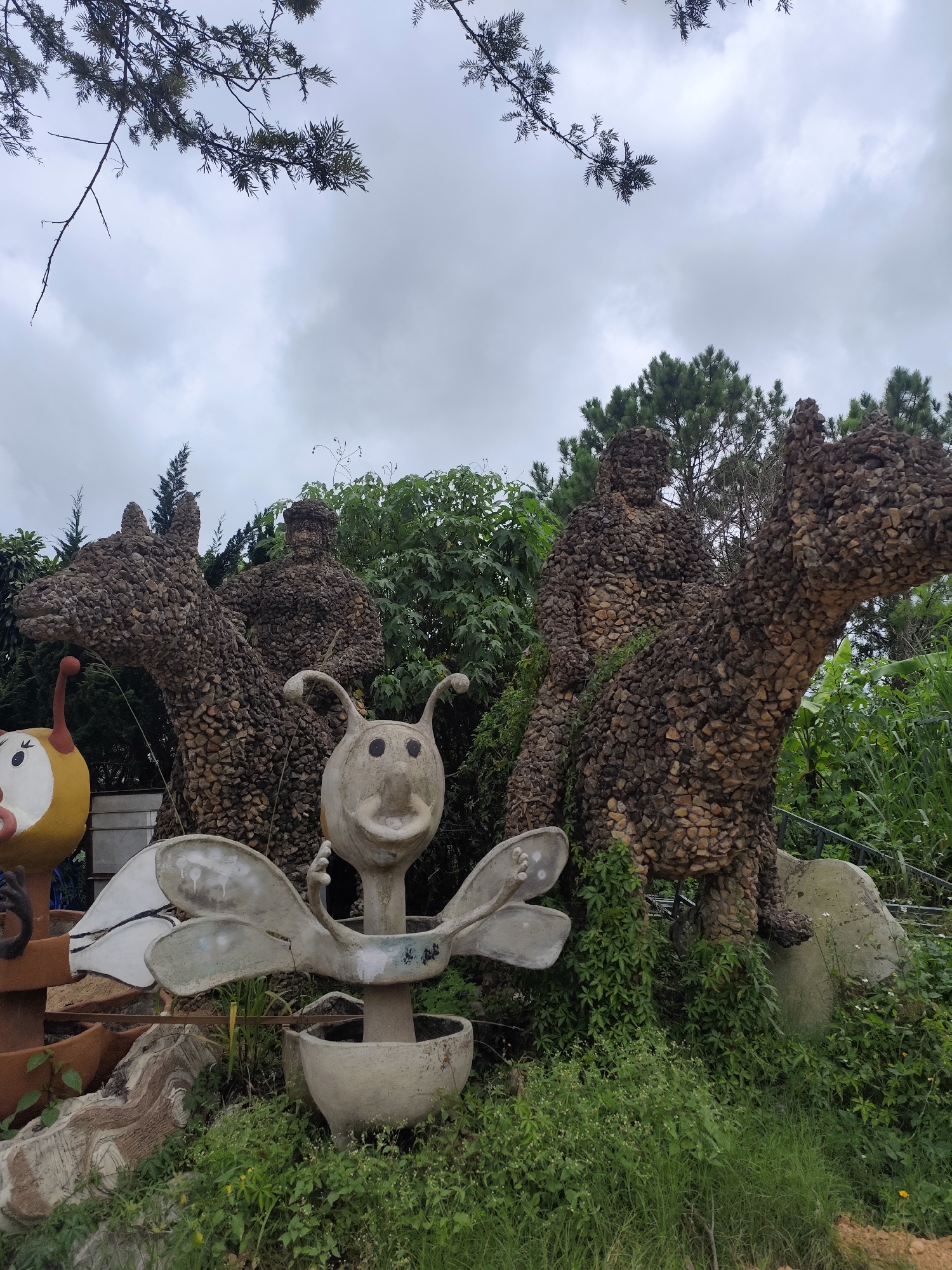 The image depicts an outdoor sculpture garden in Lạc Dương, Vietnam, featuring several whimsical stone and concrete figures amidst lush greenery and a cloudy sky. In the foreground, a light gray concrete sculpture resembling an insect with large, round eyes and antennae stands on a pedestal. To its left, partially visible, is a yellow and red round sculpture with a face. Behind these, several larger sculptures are prominent. Two appear to be human figures, constructed from a mosaic of small, dark stones, seated atop large, horse-like creatures also made of stone. One of these stone horse-and-rider sculptures is on the left, and another is on the right. Vines and other plants grow around the base of the stone figures. The background consists of dense green foliage and evergreen trees, with a overcast sky overhead. The overall scene suggests a public art installation or a garden attraction.