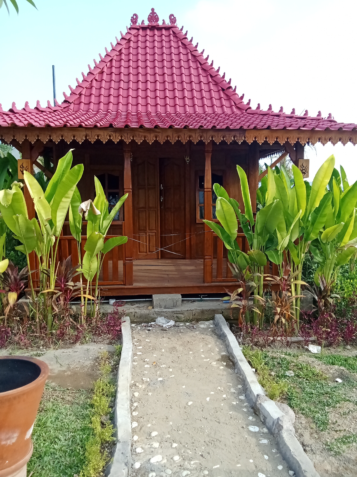 This image captures a traditional Indonesian house with a vibrant red, tiered roof, situated in a tropical garden setting. The house is constructed primarily of dark wood, featuring ornate carvings along the eaves and a welcoming wooden porch. Large, lush green leaves of what appear to be banana plants or similar foliage flank the entrance, adding a tropical feel to the scene. A sandy pathway, bordered by concrete curbs, leads towards the house, with small pebbles scattered along its surface. In the foreground, a large, terracotta pot is partially visible on the left. The background shows a clear sky. The architectural style and lush vegetation suggest this location is within Selemadeg Timur, Bali, Indonesia, a region known for its traditional Balinese houses and abundant natural beauty.