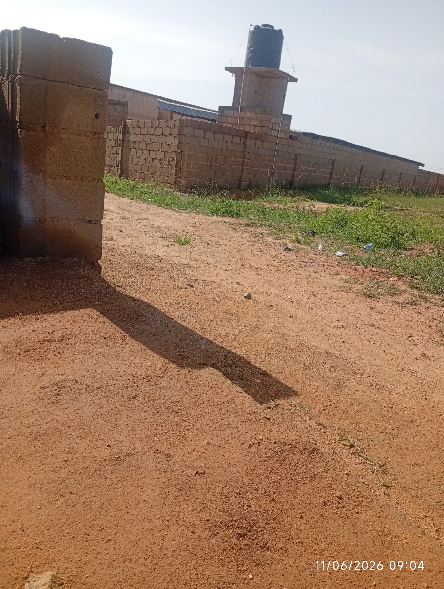 The image shows an outdoor scene in DU, Nigeria, on what appears to be a construction site or an undeveloped area. In the foreground, a dirt path is visible with a long shadow cast by a structure on the left. The path is reddish-brown in color and appears dry and dusty.
To the right of the path, there is a patch of green grass with some scattered debris, including plastic bottles and other litter. In the background, a long brick wall stretches across the frame, forming a boundary. Above and behind the wall, a large black water tank is mounted on a concrete platform, supported by a brick structure. The sky is clear and bright. The overall impression is of a utilitarian and somewhat neglected environment. The timestamp "11/06/2026 09:04" is visible in the bottom right corner of the image.