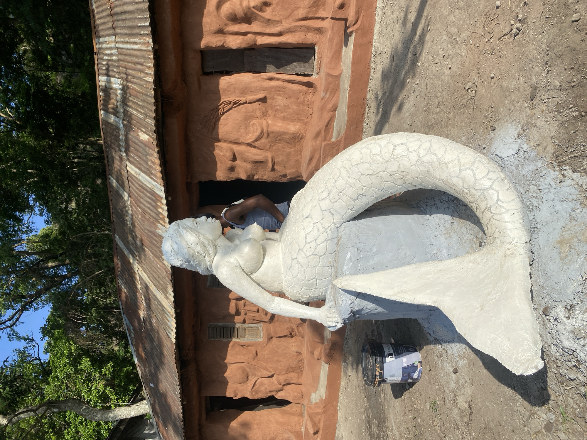An unfinished statue of a mermaid is the central subject of this outdoor scene. The statue, predominantly white with some areas painted grey, depicts a woman from the waist up, with a scaled tail extending behind her. She is seated on what appears to be a rock or pedestal. A person, wearing a grey tank top and dark shorts, is partially visible behind the statue, seemingly working on it or resting. The mermaid statue is positioned in front of a rustic building with a corrugated metal roof and textured, reddish-brown walls that resemble rock formations. The building has dark openings that suggest doorways or windows, with wooden slats visible in some. The ground around the statue and building is a mix of dirt and small stones. A black paint can with a label is on the ground near the statue's tail.
The scene appears to be set outdoors during the daytime, with clear blue sky visible through the lush green foliage of trees in the background. Sunlight casts shadows on the ground, suggesting it's a sunny day. The overall impression is one of an artistic endeavor in a somewhat undeveloped or natural setting, possibly a workshop or a roadside attraction. The unfinished nature of the statue and the presence of the paint can indicate ongoing work.
