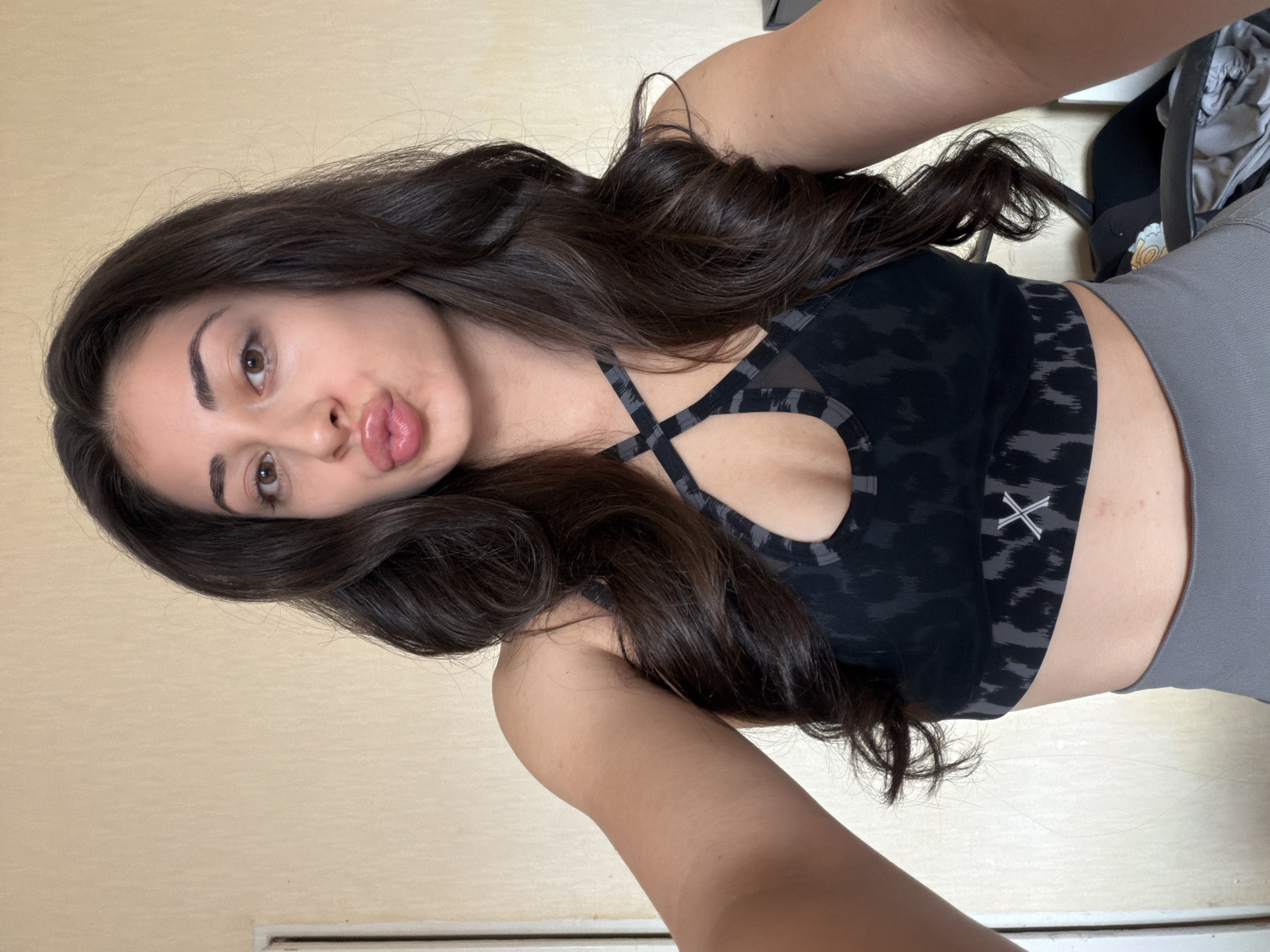 A female subject, appearing to be an adult, is depicted in a selfie, posing with pursed lips in a "kissing" expression and direct eye contact with the camera. She has long, dark, wavy hair, brown eyes, and groomed eyebrows. She is wearing a dark grey and black leopard-print sports bra featuring a criss-cross strap design at the neckline and a white 'X' logo on the band, along with light grey athletic leggings. Her extended arms indicate she is holding the camera. The indoor background consists of a plain, light beige-colored wall. A white door frame is partially visible on the far left. On the far right, a dark object, possibly a chair, with some grey fabric, is partially visible. The specific location within Sofia, Bulgaria, cannot be determined from the visual information in the image.