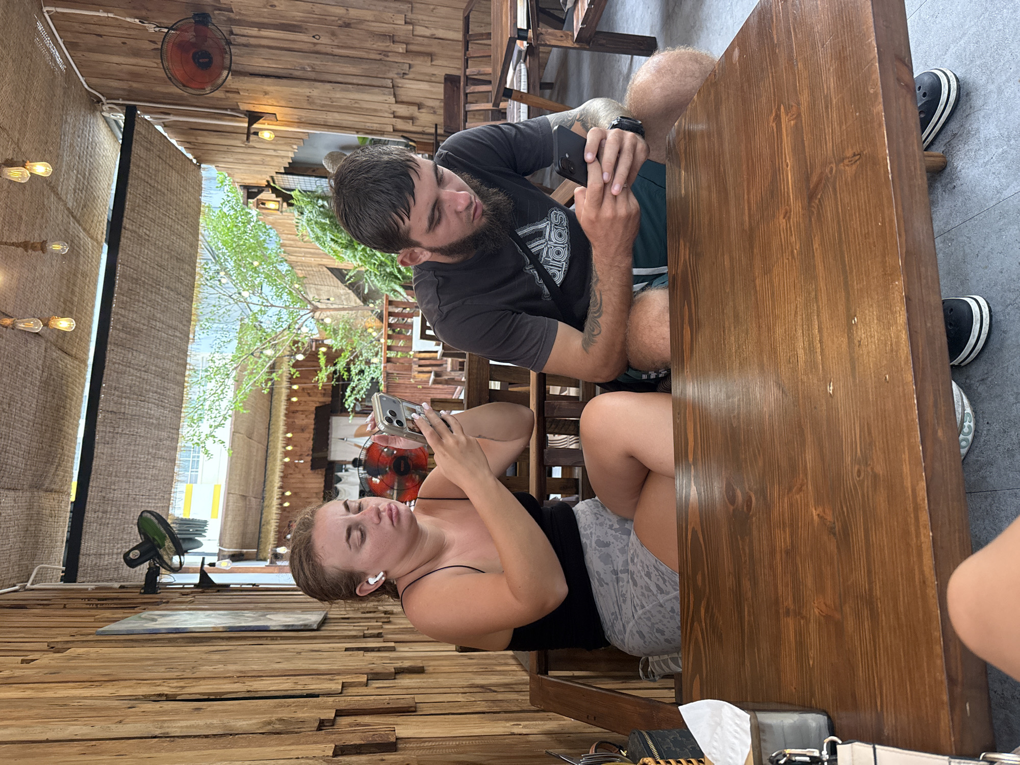 Two individuals, a woman and a man, are seated at a wooden table in an outdoor cafe with a bamboo-covered ceiling. The woman, on the left, wears a black tank top and patterned shorts, and has white earbuds in her ears. She is looking down at her phone, which she is holding in her right hand. The man, on the right, has a beard and is wearing a dark t-shirt and dark shorts. He is also looking down at his phone, which he is holding in both hands. The cafe has wooden walls and is decorated with string lights and fans. Plants are visible in the background. The scene appears to be in the city of Nha Trang, Vietnam.