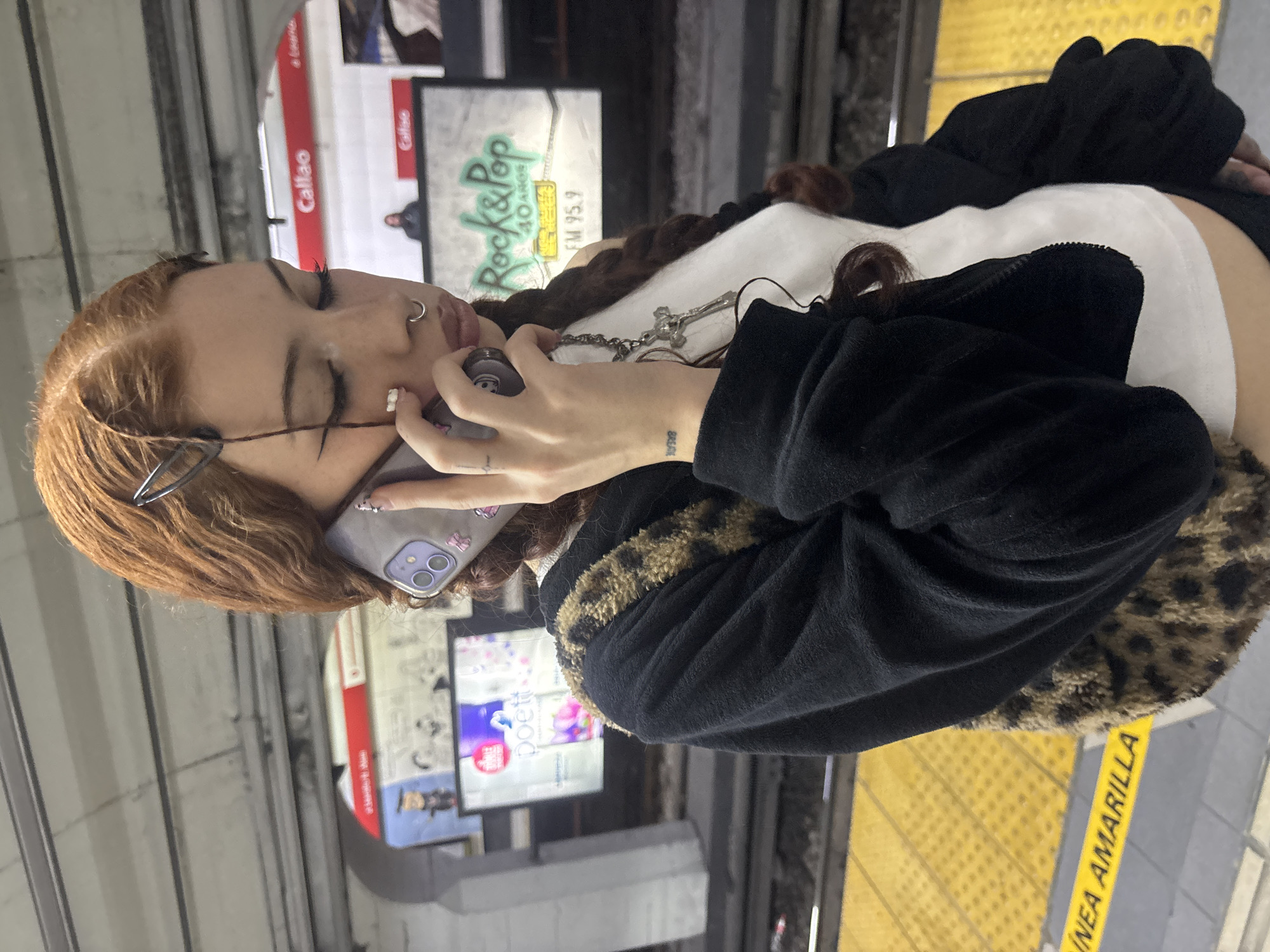 A young adult female stands on a subway platform. She has brown-red hair, multiple facial piercings, and glasses hanging from her ear. She is wearing a white top under a black, leopard-trimmed jacket, and a silver cross necklace. She is holding a light purple iPhone to her face. Subway advertisements are behind her on the wall. The subway platform has a yellow stripe with "LINEA AMARILLA" printed on it, indicating that it is in the city of Buenos Aires, Argentina.
