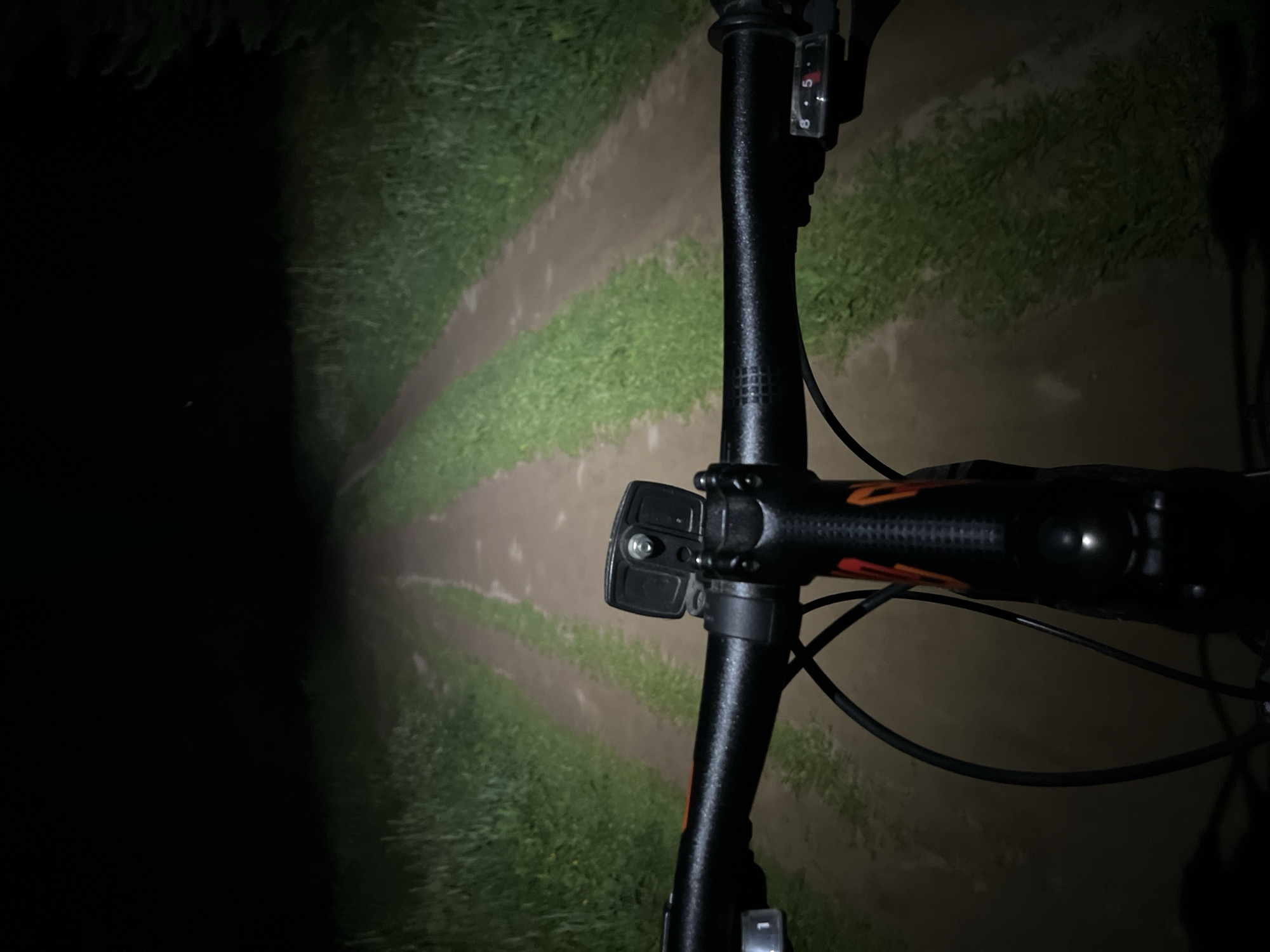 The image depicts a first-person perspective of a person riding a bicycle at night on a dirt road. The bicycle's headlight illuminates the path ahead, which is flanked by grass. The handlebars and front of the bicycle are visible in the foreground. The road appears to be in a rural or semi-rural area, possibly on the outskirts of Chaikovsky, Russia, given the context provided, though the image itself does not contain specific geographical markers to confirm this location definitively. The scene is dark, with the only significant illumination coming from the bicycle's light. There are no other people or vehicles visible.