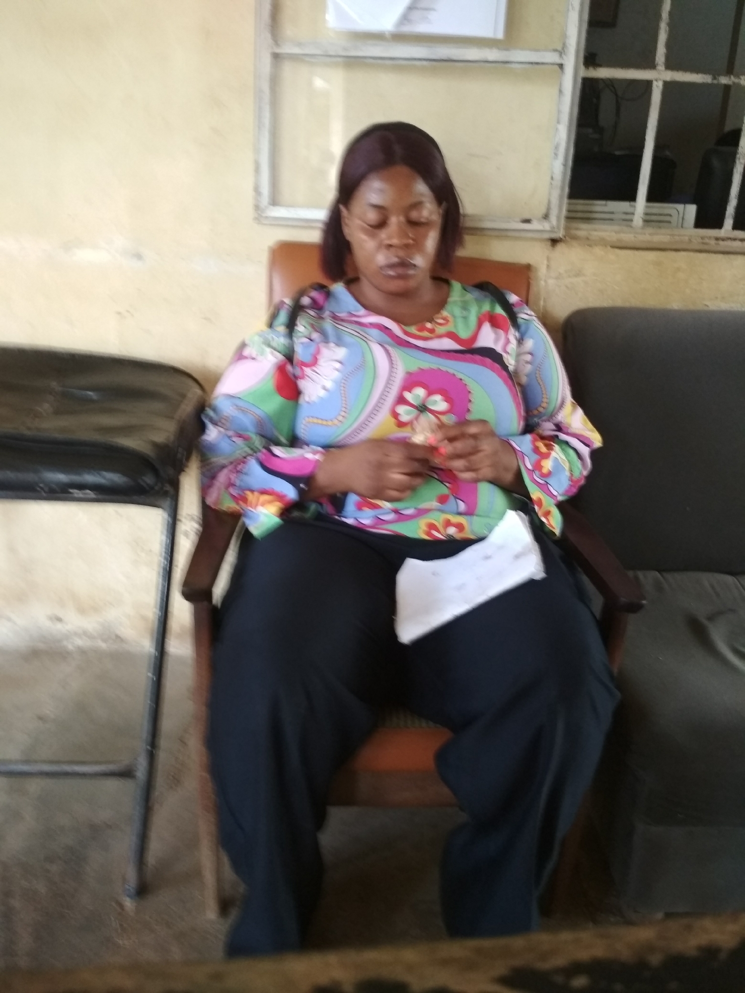 A Black woman with dark hair pulled back is sitting in a chair with her eyes closed. She is wearing a colorful, long-sleeved blouse with a paisley pattern in shades of pink, blue, green, and yellow, and loose black pants. She is holding a small, folded piece of white paper in her hands, and another piece of paper is resting on her lap. She appears to be in an indoor waiting area. To her left, there is a dark, metal chair with a worn black seat. To her right, there is a dark gray upholstered bench. The wall behind her is painted a light yellow. A window with a white frame and metal bars is visible in the background. The floor is a dull concrete. The scene depicts a person waiting, possibly in a public facility.