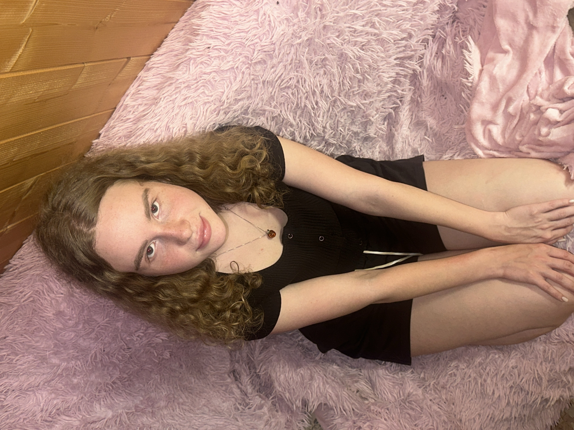 A young woman with long, curly brown hair, fair skin, and freckles is depicted lying down on a soft, fluffy light purple blanket. She is wearing a black short-sleeved top with buttons and a silver necklace featuring a reddish-brown pendant. Her fingernails are painted a light color, possibly white or pale pink. She lies with her head tilted to the left, looking directly at the camera with a gentle, slight smile. Her knees are drawn up, and her arms are crossed, resting on her upper legs. To the left and in the upper background, a wooden wall with horizontal planks is visible. Another lighter pink, smoother blanket or textile can be seen on the right side, adjacent to the purple one. The setting appears to be indoors, with soft, even lighting, suggesting an intimate and relaxed atmosphere, possibly in a bedroom or living area. No text is visible in the image. The location context provided is Vasilyevo, Russia.