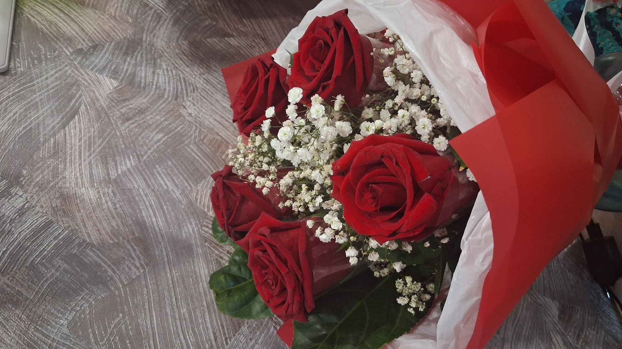 A bouquet of red roses and baby's breath is displayed against a textured gray background. The roses are vibrant and velvety, with some appearing fully bloomed and others in various stages of opening. Delicate white flowers of baby's breath are interspersed throughout the bouquet, adding a light and airy contrast to the deep red of the roses. The bouquet is partially wrapped in white paper and red paper, with the red paper forming a prominent, curved element in the foreground, suggesting it's part of the wrapping or a decorative accent. The background is a textured wall, possibly wallpaper or a painted surface, with visible brushstrokes or a patterned design that creates a sense of depth and movement. The lighting appears to be soft, illuminating the flowers and highlighting their rich colors and intricate details. The overall impression is one of elegance and classic beauty. The image does not provide any information about its location in Koltsovo, Russia, or any people or actions.