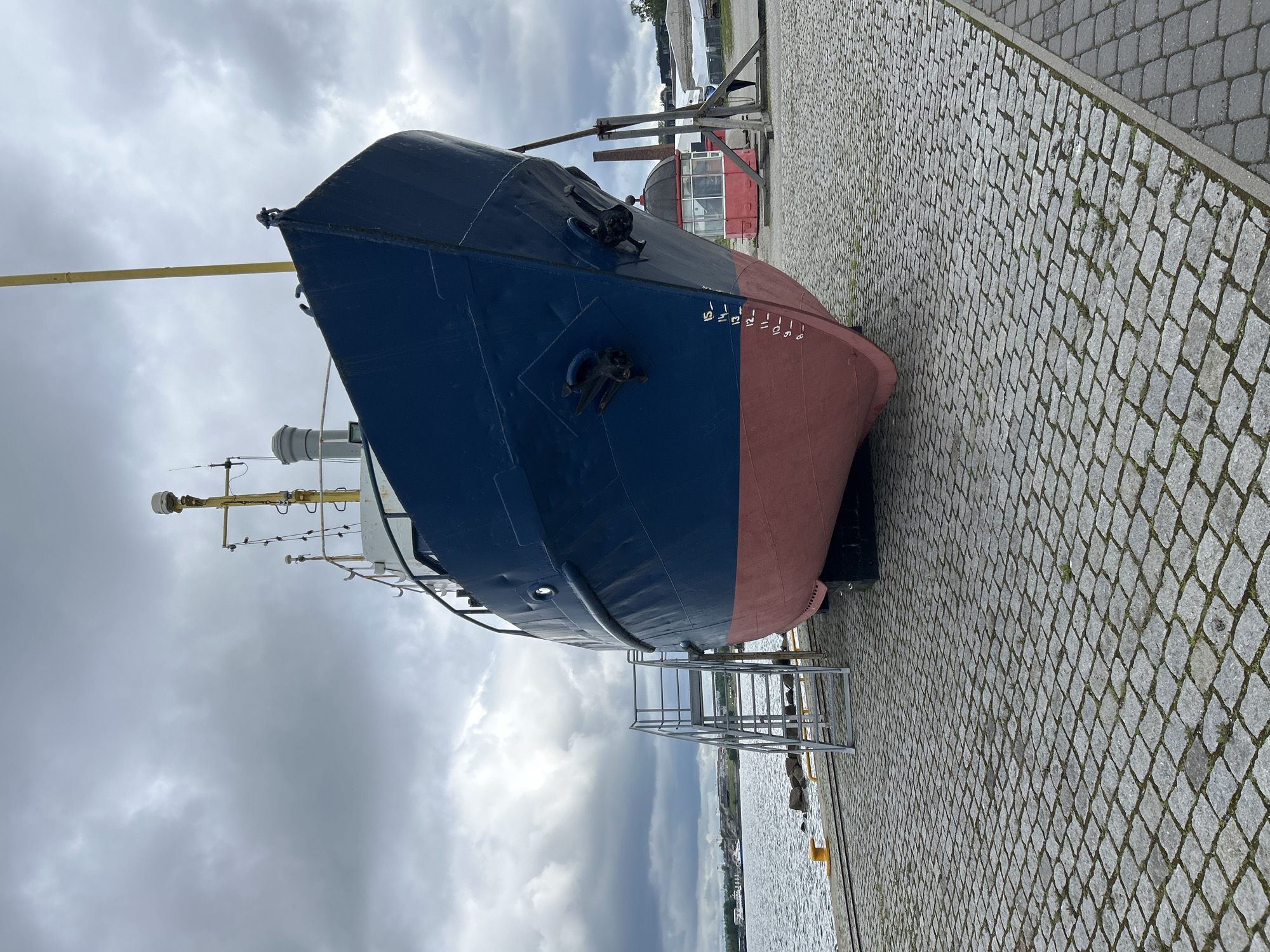 A blue and red ship is parked on a cobblestone pier in Tallinn, Estonia. The ship is oriented towards the camera, with its bow slightly to the right. A set of metal stairs leads up to the ship's deck on the left side. The water is visible on the left, with a yellow buoy in the foreground. The sky is cloudy. In the background, there are some buildings and a red structure with a glass window. The ship has anchors visible on its bow. Depth markings are painted on the hull.