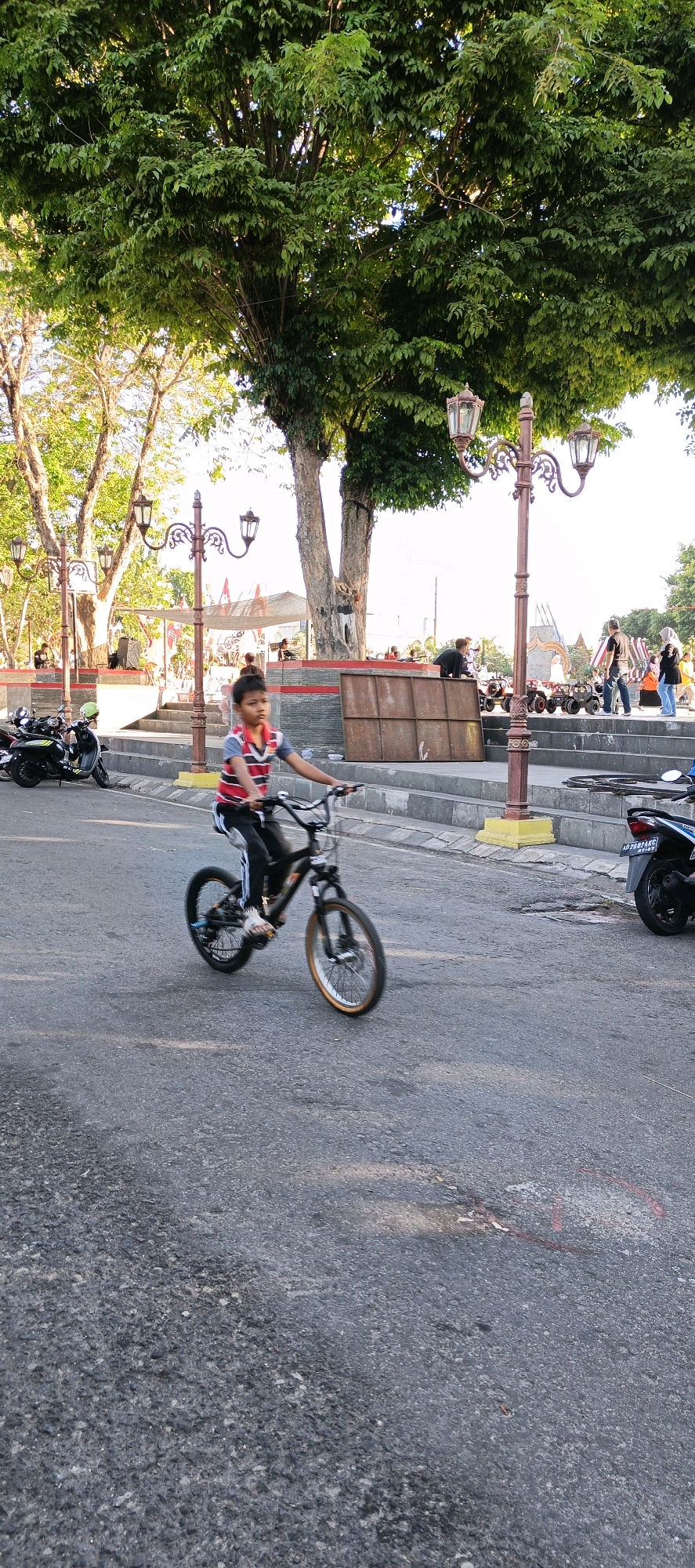 The image depicts a child riding a bicycle on a paved street in front of a park or public square in Klaten, Indonesia. The child, wearing a striped shirt and dark pants, is captured in motion, with the bicycle wheels blurred. In the background, there are trees, lampposts, a set of stairs, and several people, some of whom appear to be sitting or standing. Motorbikes are parked on the left side of the street. The overall scene suggests a sunny day in an urban public space.
