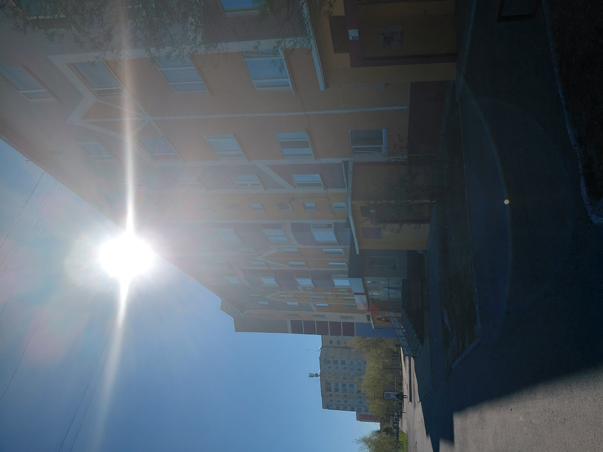 The image, taken in Nadym, Russia, depicts a bright, sunny daytime street scene dominated by a multi-story building. This building, on the left side of the frame, is painted in warm yellow and orange tones, featuring numerous rectangular windows and distinctive white geometric lines that outline sections of its facade. The sun is intensely bright in the upper left corner, casting a strong glare and lens flare across the clear blue sky and the upper portion of the building, making some details hazy.
To the right, a paved street or sidewalk stretches into the distance, bordered by patches of green grass and some sparse trees. Further in the background, another multi-story building, lighter in color (possibly grey or beige) and also featuring many windows, is visible with an antenna on its roof. A small, dark figure, likely a person, can be discerned near this distant building on the right side of the road.
The clear, bright sky indicates sunny weather, and the high sun position suggests it is likely mid-morning or afternoon. The architecture of the buildings, with their block-like design, is typical of urban developments in regions like Nadym. The scene is static, showing no specific activity or event beyond a general street view. Small signs are visible on the main building, but any text on them is illegible due to the distance and image resolution.