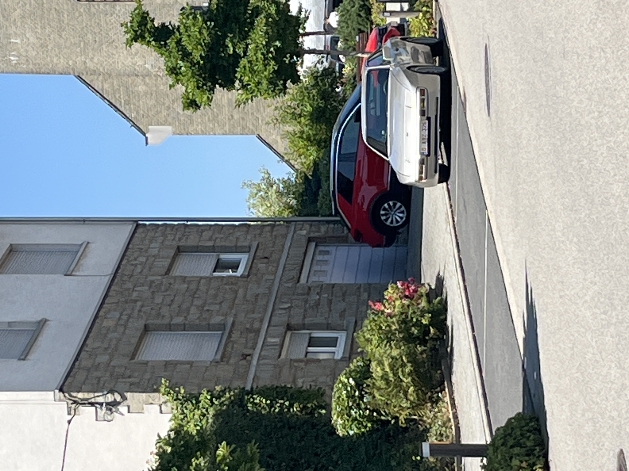 The image shows a street scene in Saint Vith, Belgium. In the foreground, a light-colored, angular sports car is parked on the right side of the road. Behind it, a red SUV is parked. The cars are on a paved surface next to a sidewalk and a small patch of grass with bushes and some pink flowers. To the left, a multi-story building with a stone facade and white upper floors is visible, featuring windows with blinds and a garage door. To the right, another building with a stone facade rises, with a large green tree in front of it. The sky is clear and blue. There are no people clearly visible in the image.