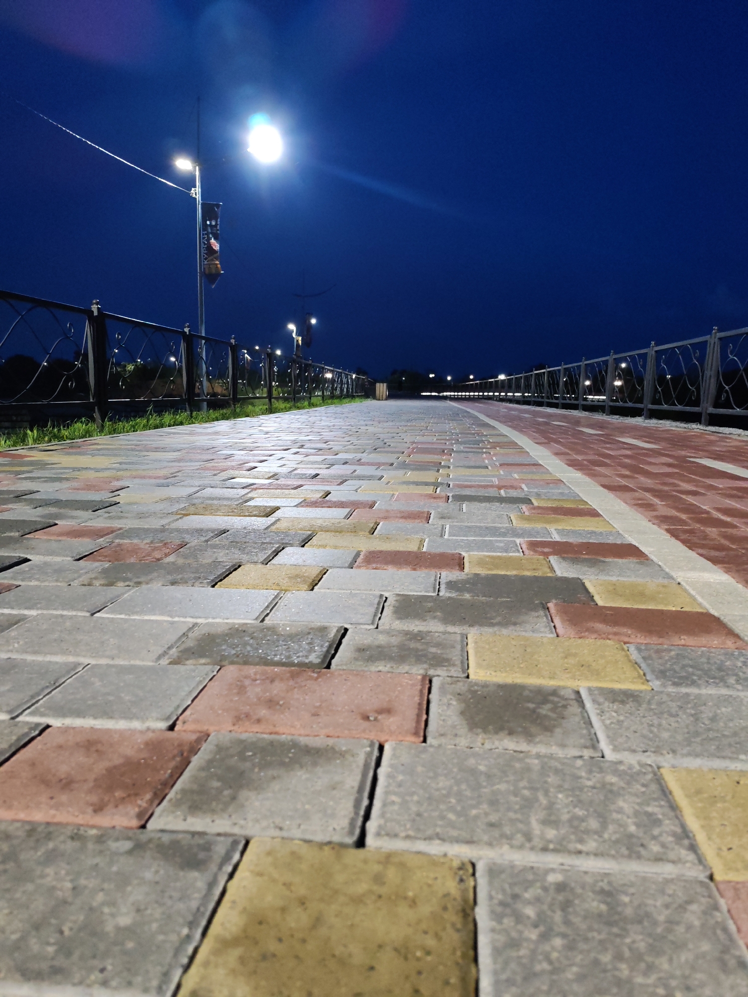 A low-angle shot captures a wide, paved walkway at dusk. The walkway is constructed from rectangular interlocking pavers in a variety of colors, including grey, reddish-brown, and yellow. The pavers on the left are wet, with visible reflections of the lights. A black metal fence with an ornate pattern runs along the right side of the walkway, separating it from a strip of red pavers and then a grassy area. In the background, several streetlights illuminate the scene, casting a bright glow against the deep blue twilight sky. One streetlight in the foreground is particularly bright and has a banner hanging from it. The walkway recedes into the distance, creating a sense of depth and perspective. The overall scene is serene and well-lit, suggesting a public space that is maintained and inviting.