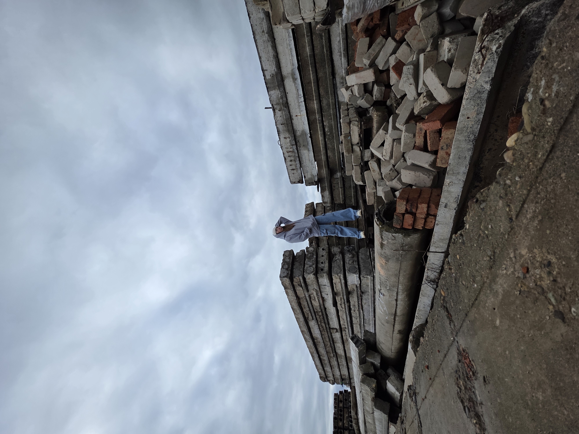 An image, rotated on its side, shows a person standing on a tall stack of weathered concrete slabs. The individual, dressed in a grey hooded sweatshirt, light blue jeans, and white sneakers, is positioned towards the center-left of the frame, looking up at the cloudy sky with one hand raised towards their head.
The scene depicts an outdoor, industrial or construction-related environment. The concrete slabs beneath the person are rectangular, thick, and appear worn. To the right of these slabs, and extending further into the background, are large, haphazard piles of bricks, a mix of traditional red and lighter grey or white types. A rough, textured concrete wall or surface frames the right side of the image, suggesting a larger structure or boundary.
The sky above is overcast and grey, indicating a cloudy daytime setting without direct sunlight. The overall impression is one of a material storage yard, a demolition site, or an abandoned industrial area. There is no discernible text visible in the image. The person's activity seems to be posing or observing the surroundings from an elevated position.
