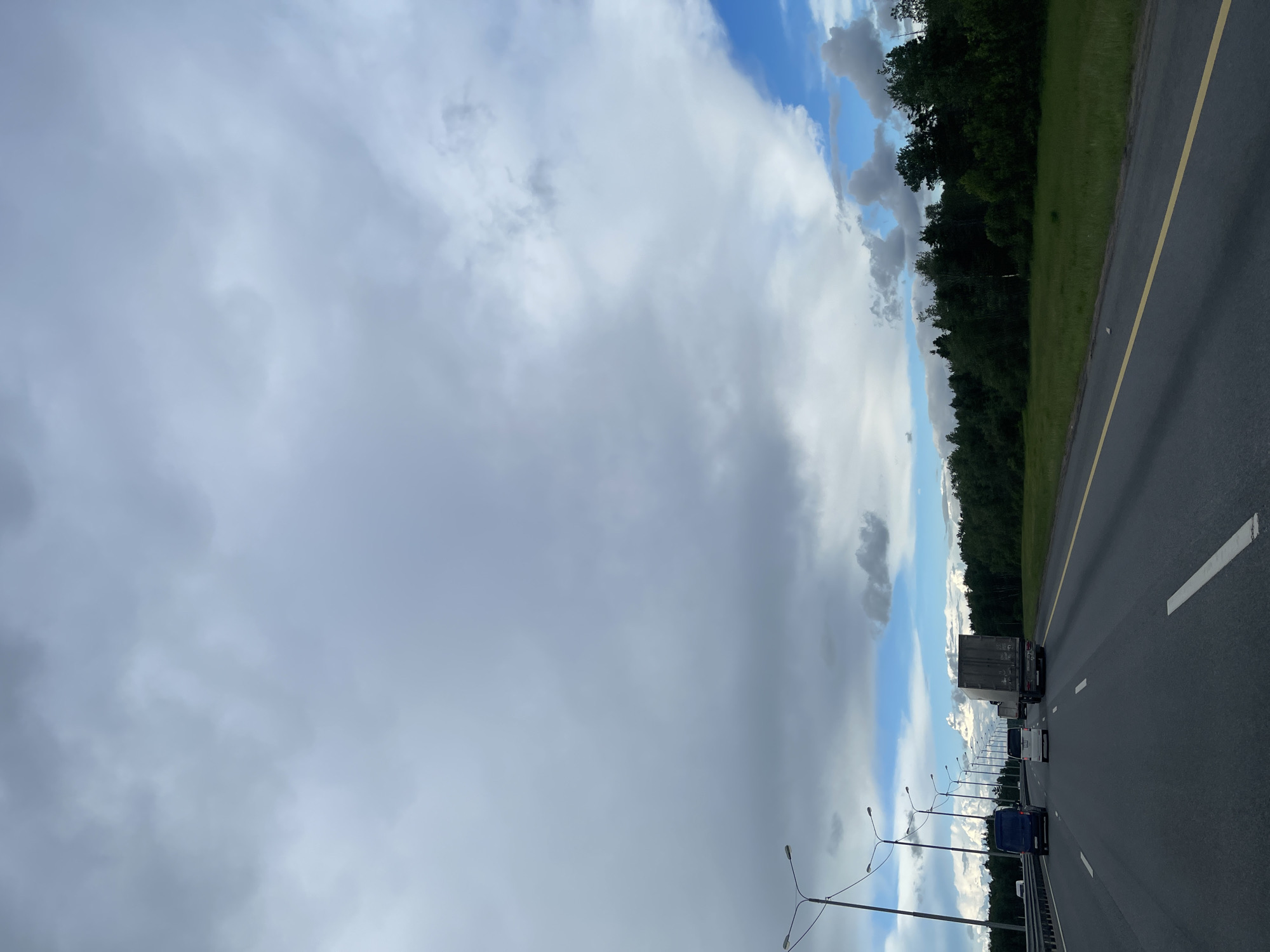 The image shows a view from a vehicle driving on a highway near Бавыкино, Russia. The sky is dominated by heavy cloud cover, with patches of blue visible. To the right of the road, there is a line of trees bordering a grassy area. The road is asphalt with white lane markers and a yellow line on the right edge. A line of streetlights runs along the side of the highway. Several vehicles, including a semi-truck and smaller cars, are visible on the road ahead.