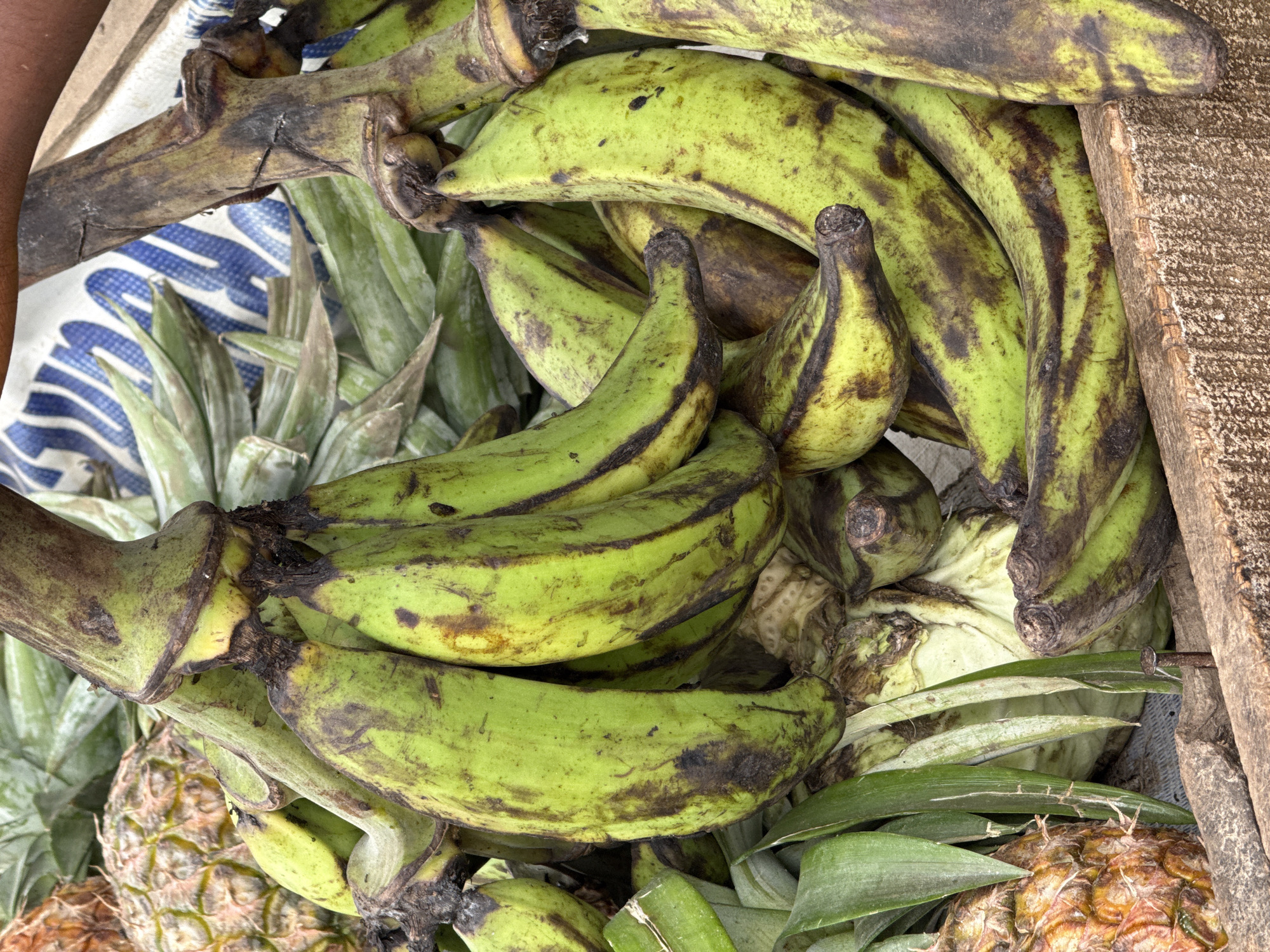 A close-up, eye-level shot captures a cluster of green plantains in a rustic wooden crate. The plantains, varying in ripeness with some showing brown splotches, are piled together, with their stems and some leaves visible. In the background, a pineapple is partially visible, along with a blue and white patterned sack and more green leaves. The lighting is natural and appears to be daytime. The scene suggests a market stall in Abuja, Nigeria, focusing on fresh produce.