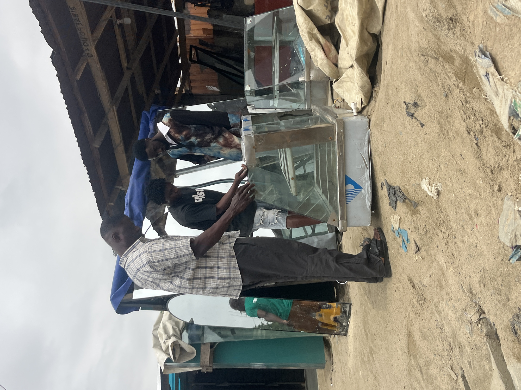 In Ado, Nigeria, three men are gathered around two glass display cases on a sandy lot. The man on the left, wearing a plaid shirt and dark pants, appears to be supervising. The man in the middle, in a black t-shirt and denim shorts, is looking down at the display case. The man on the right, in a colorful patterned shirt, is also looking at the display case. The background shows a makeshift shelter with a corrugated metal roof, and several large mirrors are propped up behind the men. The scene suggests a commercial setting, possibly a market or a repair shop for display cases. The ground is covered in sand, with some debris scattered around. The sky is overcast, indicating a cloudy day.