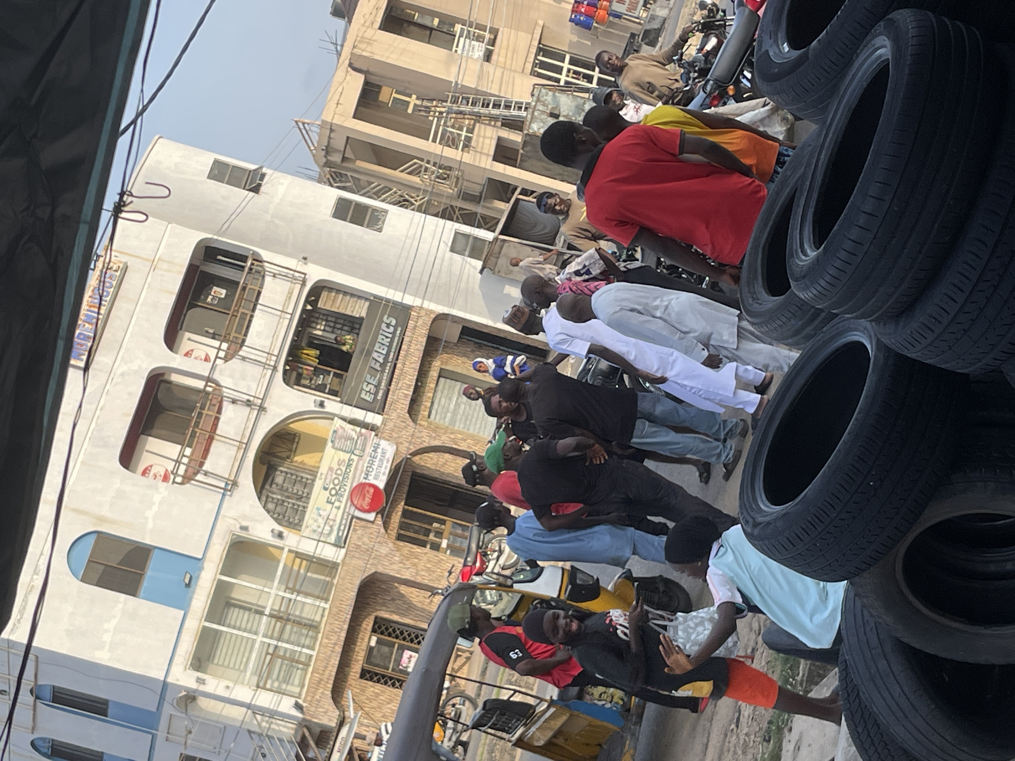 This media file captures a street scene with a group of people gathered around vehicles and stacks of tires. The image is oriented vertically, suggesting it might have been taken with a phone. In the foreground, several black tires are piled high, some appearing new and others worn. Behind the tires, a yellow and white three-wheeled vehicle, possibly an auto-rickshaw, is visible, with people interacting around it.
A prominent building with multiple floors forms the backdrop. The architecture suggests a commercial area, with shops or businesses on the ground floor. Signs on the building include "MICO," "FOODS & PROVISIONS," "MOREMI COLLEGE RESTAURANT," and "ESE FABRICS," indicating the presence of various establishments. The Coca-Cola logo is also visible on one of the signs.
Several individuals are present, appearing to be engaged in various activities, such as loading or unloading items, conversing, or simply standing around. They are dressed in casual clothing, with some wearing t-shirts and others more traditional attire. The overall atmosphere seems to be that of a bustling marketplace or a busy street with commercial activity. The time of day appears to be daytime, with bright lighting, and the weather seems clear. Some people are looking towards the camera, while others are focused on their immediate surroundings. The scene depicts a lively, everyday moment in what appears to be an urban environment in an African country, given the context provided by the text on the signs.