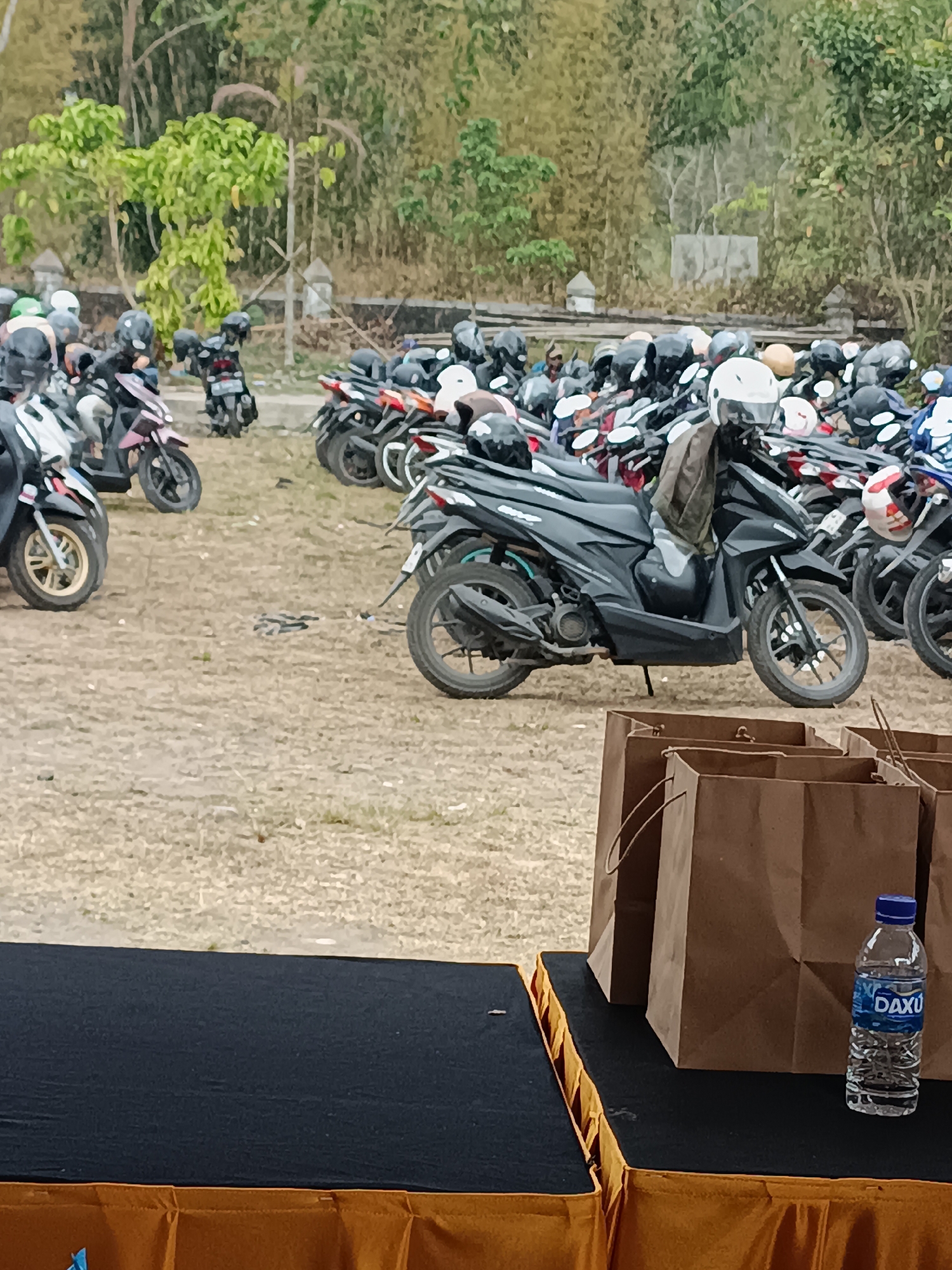 The image captures an outdoor scene in Hargobinangun, Indonesia, featuring a large number of parked motorbikes. The motorbikes, primarily scooters, are arranged closely together in a gravel lot. Many of the bikes have helmets placed on their seats or handlebars. In the foreground, a black table is draped with a dark blue cloth, and an orange fabric is visible underneath. On the table sits a bottle of "DAXU" bottled water. Behind the table, several brown paper bags with handles are stacked. The background consists of a dense area of trees and foliage, with some evidence of stone structures or walls. The overall impression is that of a parking area, possibly for an event or a popular local destination.