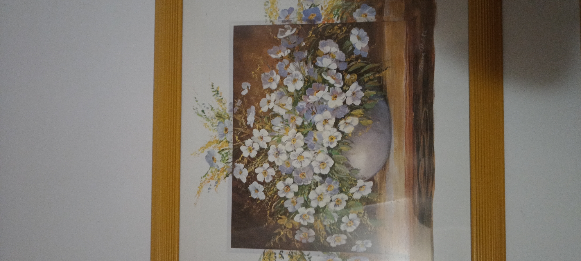 The image shows a framed painting of a floral arrangement. The painting depicts a vase filled with white and pale blue flowers, with some greenery and tall, wispy stalks of wheat or grass. The vase is light gray and appears to be made of ceramic or porcelain. It sits on a wooden surface with a dark, textured grain. The background of the painting is a muted brown and green, suggesting an indoor or rustic outdoor setting. There is a signature in the lower right corner of the painting that appears to read "Anna Pett". The painting is enclosed in a simple yellow frame. The image is taken from a slight angle, showing the painting on a wall with a white background. There is no indication of the city of Świnoujście, Poland, or any specific location within it.