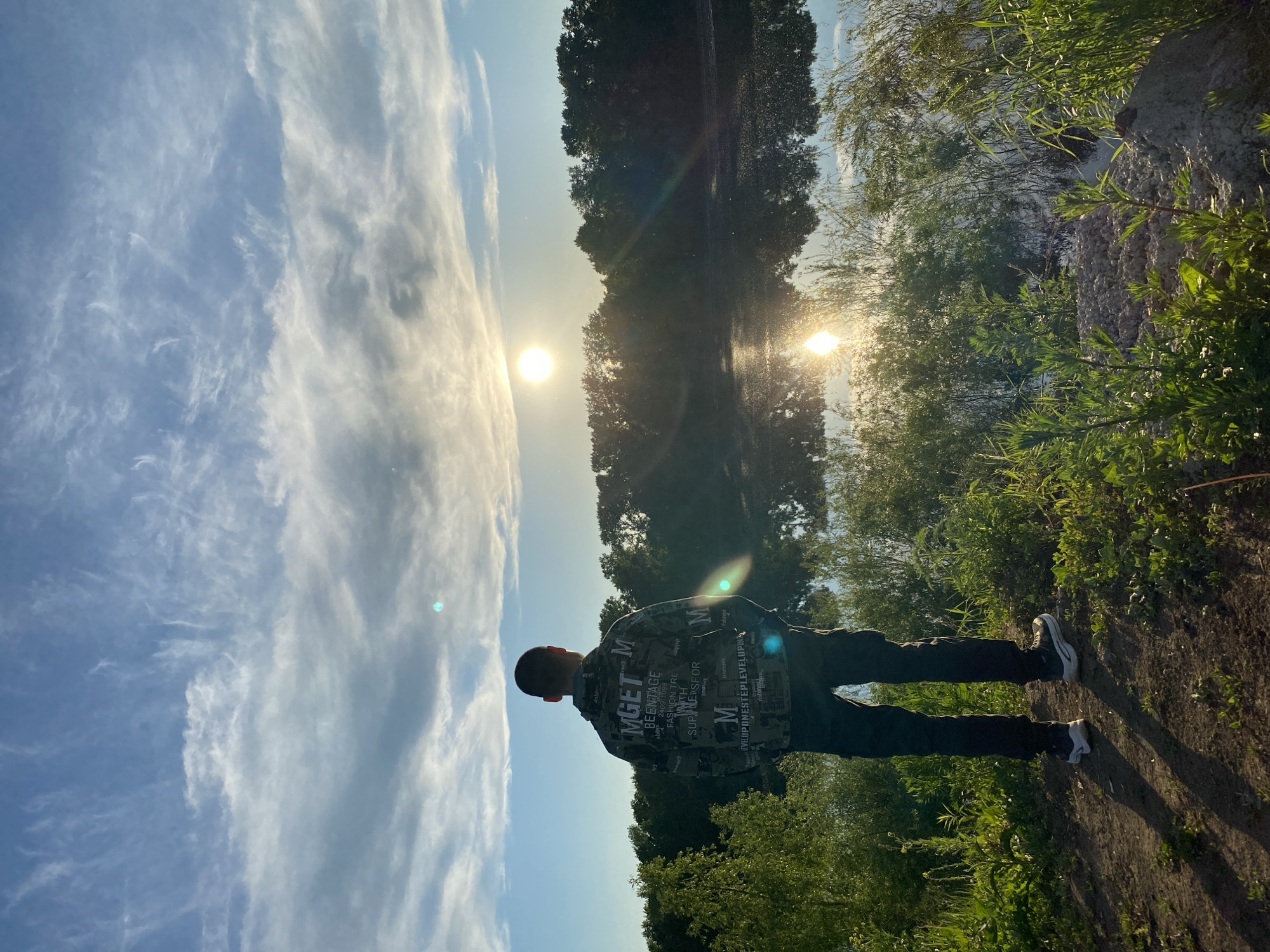The original image, correctly oriented, depicts a solitary person standing outdoors with their back to the viewer, gazing towards a bright sun setting or rising over a body of water. The scene, located in Chaikovsky, Russia, showcases a natural, verdant landscape under a partially cloudy sky.
The individual, appearing to be a male, is dressed in a dark, patterned jacket or shirt, dark trousers, and sneakers. The jacket features various textual elements as part of its design, including "MGET M," "BEENTAGE," "FASHION TRE," "SUPNERSFOR," and "EVELUPONESTEPLEVELUPINA." They also appear to be wearing red headphones. Their posture suggests a moment of contemplation or appreciation of the scenic view.
The environment is lush with green vegetation, including tall trees, bushes, and grasses that line the edge of a calm body of water, likely a river or lake. The sun, a prominent bright orb, is positioned low in the sky, casting a golden light and creating a brilliant reflection on the water's surface, strongly indicating either dawn or dusk. The sky above is a clear blue, adorned with scattered, wispy cirrus clouds. The foreground shows a dirt path or bank where the person stands. The overall impression is one of peaceful observation in a serene natural setting.