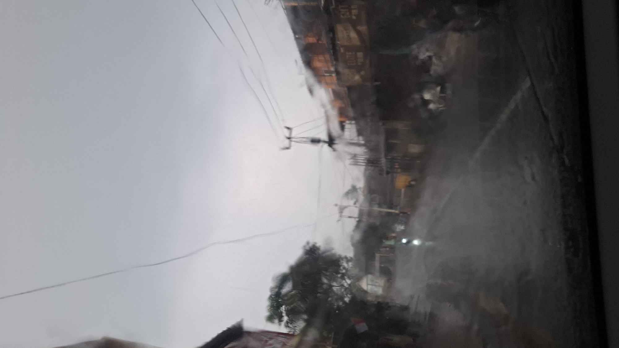 The image depicts a street scene in Cilaku, Indonesia, on a cloudy, possibly rainy day. The view is from inside a vehicle, looking out through a wet windshield, which creates a hazy and somewhat distorted effect. The road is wet and reflects the ambient light. Buildings line both sides of the street, with some appearing to be shops or residential structures. There are utility poles with wires crisscrossing the sky. A few people and what appear to be motorcycles are visible on the road, suggesting ongoing activity despite the weather. The overall atmosphere is somewhat muted and somber due to the overcast sky and wet conditions.
