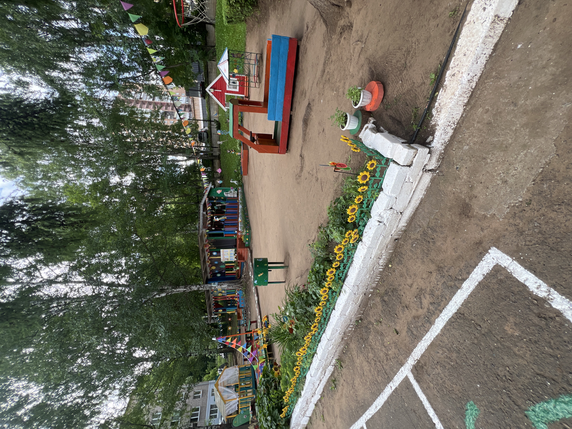 The image depicts a vibrant and colorful playground for children in Советско-Никольское, Russia. The playground is situated outdoors, surrounded by lush green trees and with residential buildings visible in the background. The ground is primarily covered with sand, a common surface for playgrounds.
Dominating the scene is a red and blue sandbox structure, designed to resemble a boat or train. Adjacent to it is a small red house with a white roof and blue trim, and a colorful canopy structure with a slide. Colorful flags are strung across the playground, adding to the festive atmosphere. A white brick border, adorned with artificial sunflowers, lines a section of the playground. Two small potted plants sit on decorative orange stands near the sunflowers.
In the foreground, there are white painted lines on the ground, possibly indicating a game area or boundary. A green metal bin is also present in the middle ground. The playground appears to be well-maintained and equipped with various play structures and decorative elements, suggesting it is a dedicated area for children's recreation. The overall impression is one of a cheerful and inviting space for outdoor play.