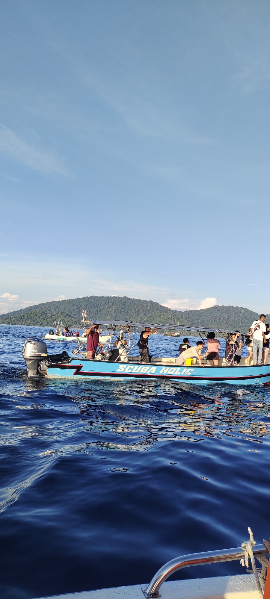 An outdoor, eye-level shot captures a bright, sunny day at sea near the city of Sabang, Indonesia. The image features a blue and white passenger boat with "SCUBA HOLIC" written in red lettering on its side. The boat is actively moving through choppy, dark blue water, creating waves that reflect the sunlight. Several people are visible on the boat, appearing to be tourists or a tour group. One person in a maroon top is standing near the front, while others are seated or standing further back. The boat has a canopy for shade and an outboard motor at the rear. In the background, a lush, green, tree-covered island with a mountainous contour rises from the sea. The sky is a clear, light blue with a few wispy white clouds. Another smaller boat is visible in the distance.
