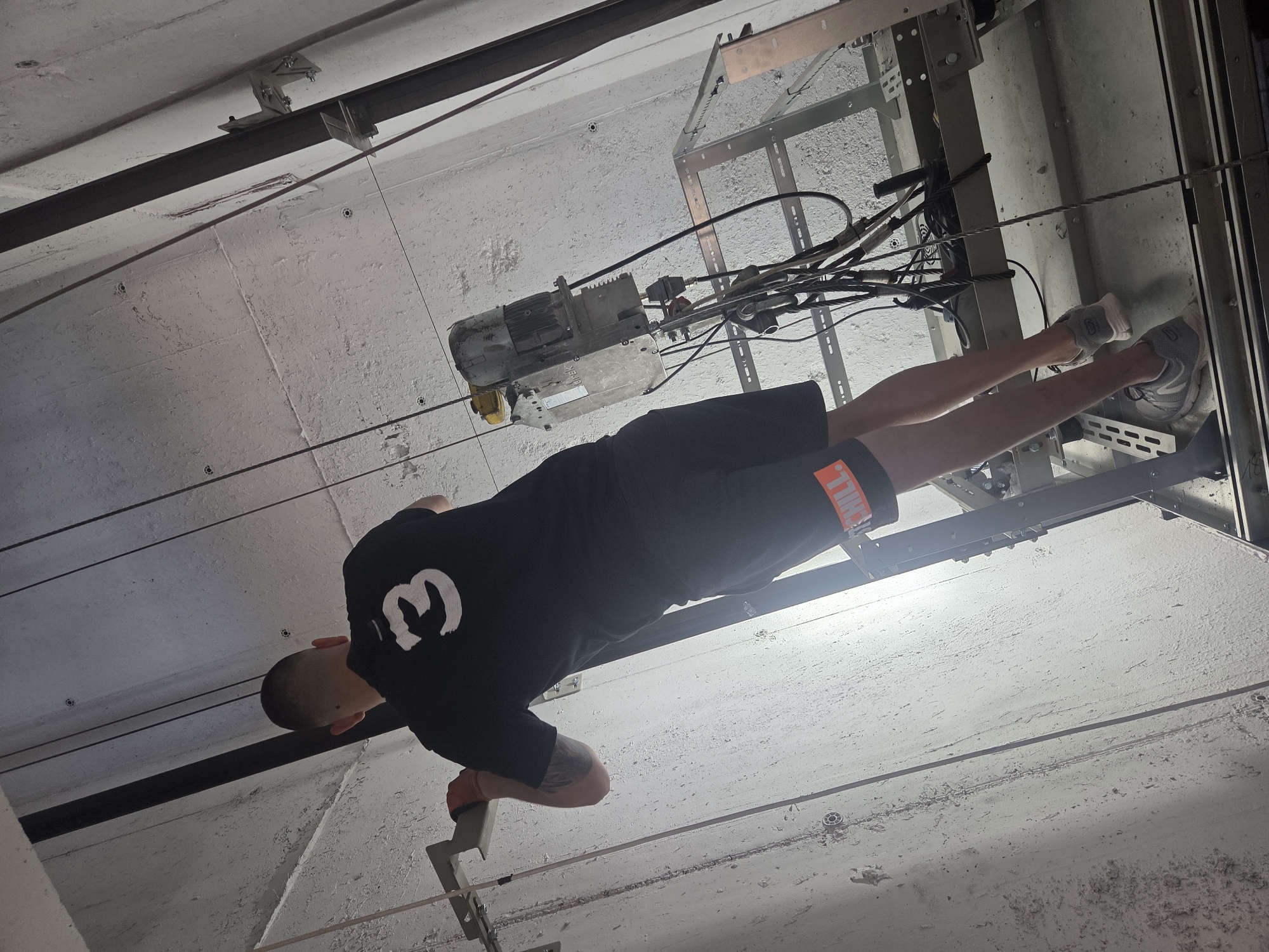 A person with short hair, wearing a black t-shirt with a white number "3" on the back and dark shorts with an orange stripe, is standing on a metal platform inside an elevator shaft. The person is facing away from the camera and appears to be working, with their right hand holding onto a metal support. The environment is industrial, with concrete walls, metal rails, wires, and an elevator motor visible. The lighting suggests it is daytime. The overall scene depicts an active construction or maintenance setting.