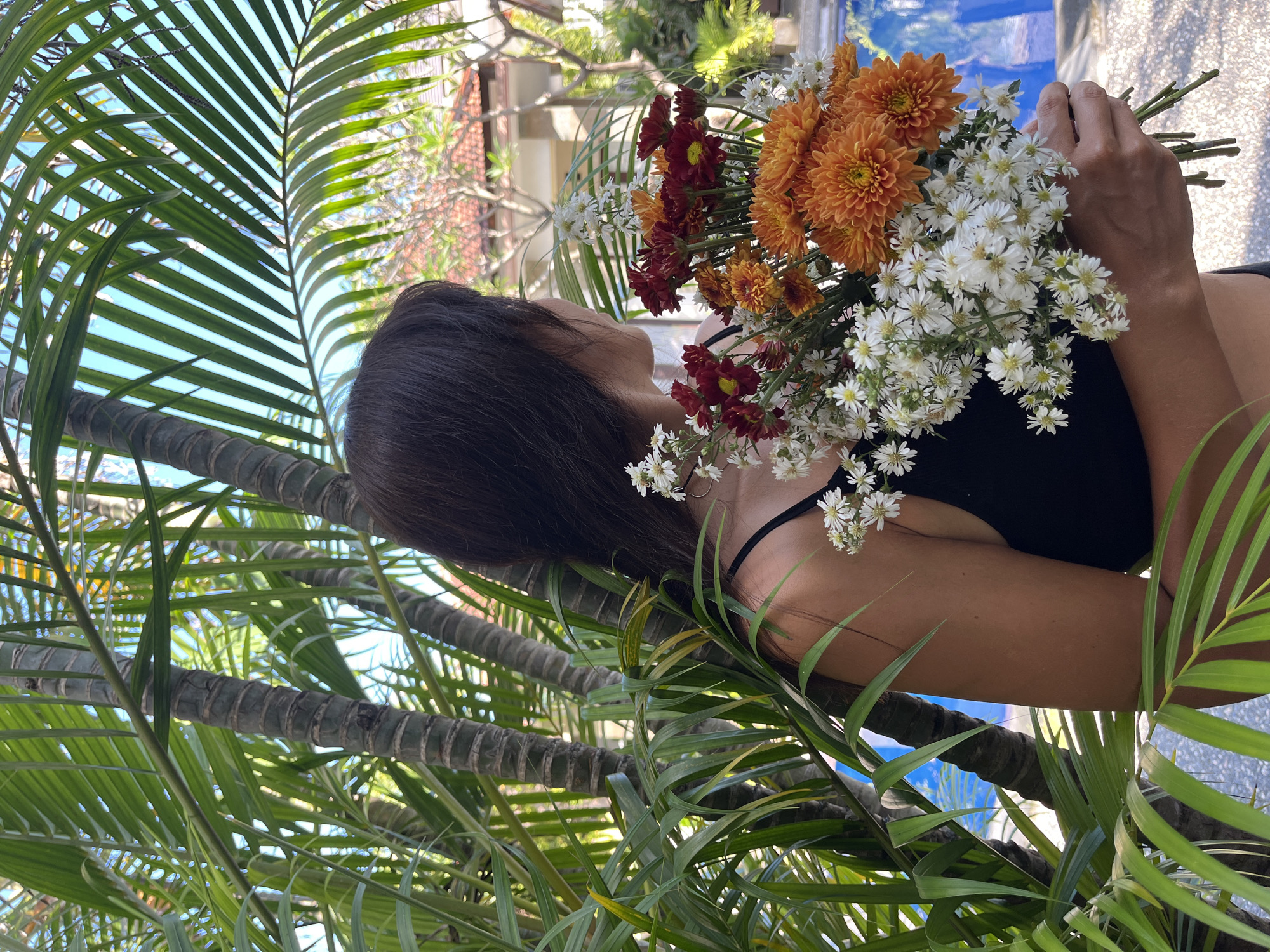 The image, oriented on its side, captures a woman amidst lush tropical foliage under bright daylight. She is seen from the left, her dark hair falling behind her, and wears a black top, possibly swimwear. She holds a large, vibrant bouquet of flowers composed of orange and deep red chrysanthemums, interspersed with small white daisy-like blossoms.
The scene is dominated by multiple green palm fronds and the textured, ringed trunks of palm trees, creating a dense, verdant foreground and midground. In the background, glimpses of a bright blue sky are visible, along with the tiled roof of a building and a blue surface, possibly a swimming pool, suggesting an outdoor resort or garden setting. The clear, strong sunlight indicates a sunny day. The composition evokes a serene, tropical atmosphere, consistent with a location like Kuta Selatan, Indonesia. There is no visible text in the image.
